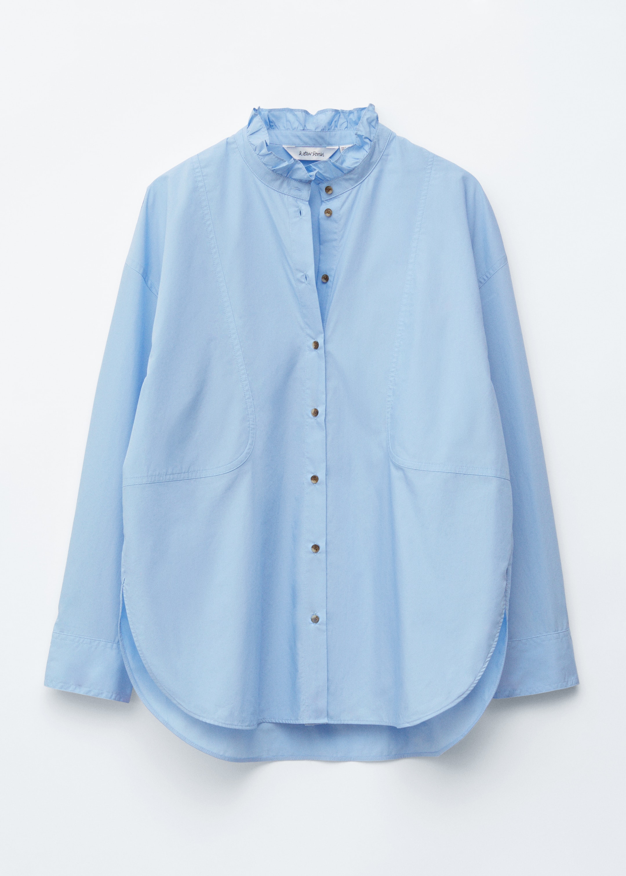 Visualizza immagine più grande: Una camicia azzurro chiaro è esposta FRONT, caratterizzata da una silhouette ampia, un colletto alla coreana con bordo arricciato e un'abbottonatura. Presenta cuciture decorative che delineano ampie aree arrotondate sulla parte inferiore anteriore ed è rifinita con un orlo arrotondato e polsini con bottoni.
