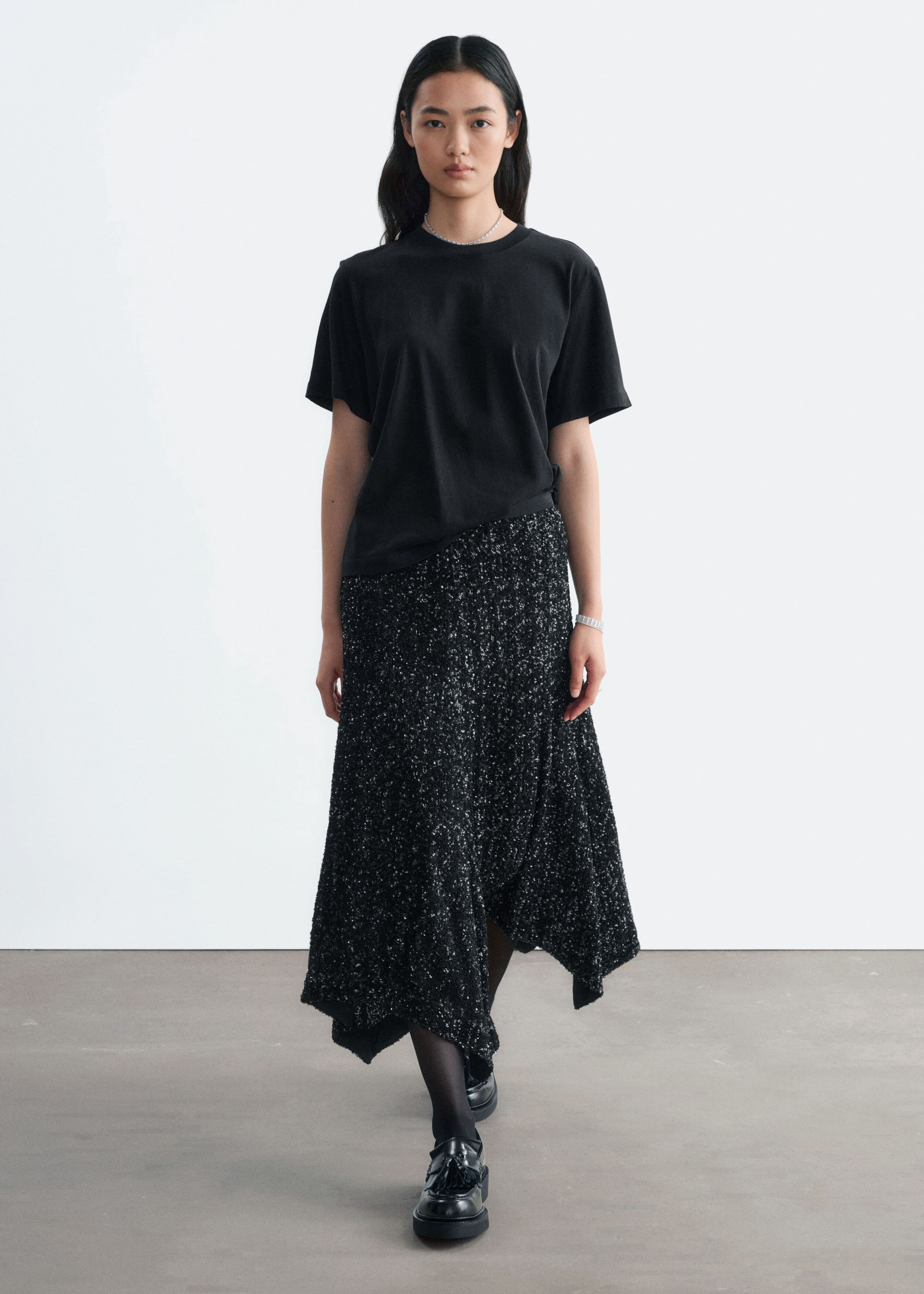 A black sequined mesh skirt featuring a textured, shimmering finish and an asymmetric hem is being worn by the model. The skirt is styled with a short-sleeved black T-shirt, black tights, and black loafers.