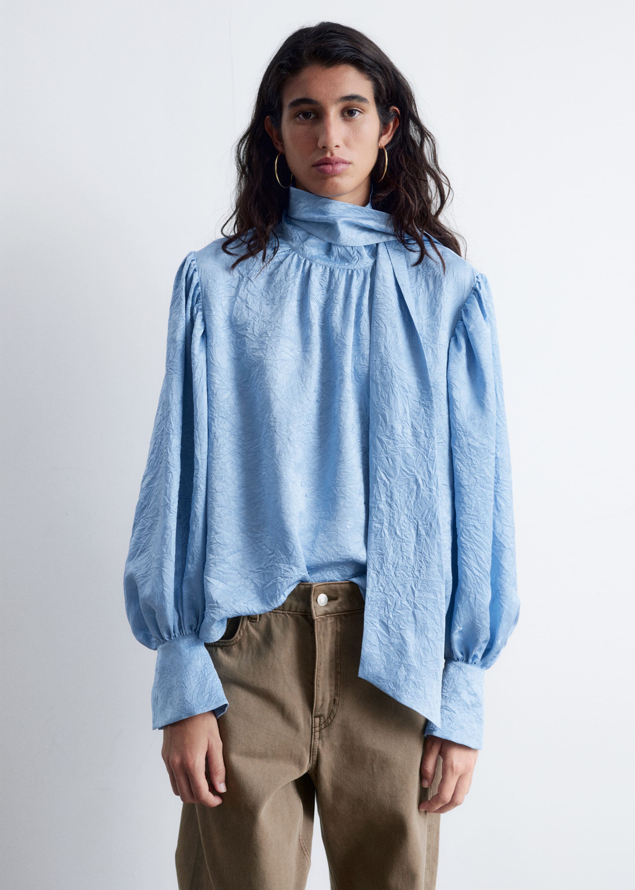 Grotere afbeelding bekijken: Een lichtblauwe gekreukelde blouse wordt gedragen, met een hoge hals en een lange strikdetail dat over de voorkant hangt. Het is ontworpen met gepofte schouders en volumineuze mouwen die bij de polsen zijn ingenomen met knoopmanchetten, en is gedeeltelijk ingestopt in een bruine broek.
