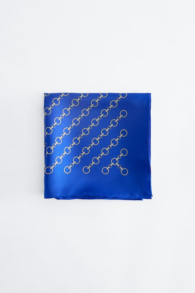 Une écharpe en soie bleu roi est pliée en carré, mettant en valeur un motif diagonal de maillons de chaîne dorés sur sa surface lisse, semblable au satin. - 1