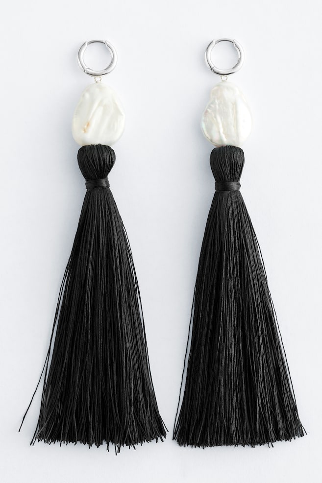 Une paire de boucles d'oreilles est présentée côte à côte, chacune arborant un anneau argenté, une perle baroque blanche de forme irrégulière et un pompon noir. Le pompon noir est suspendu à la perle, avec sa section supérieure soigneusement enroulée de fil noir. - 1