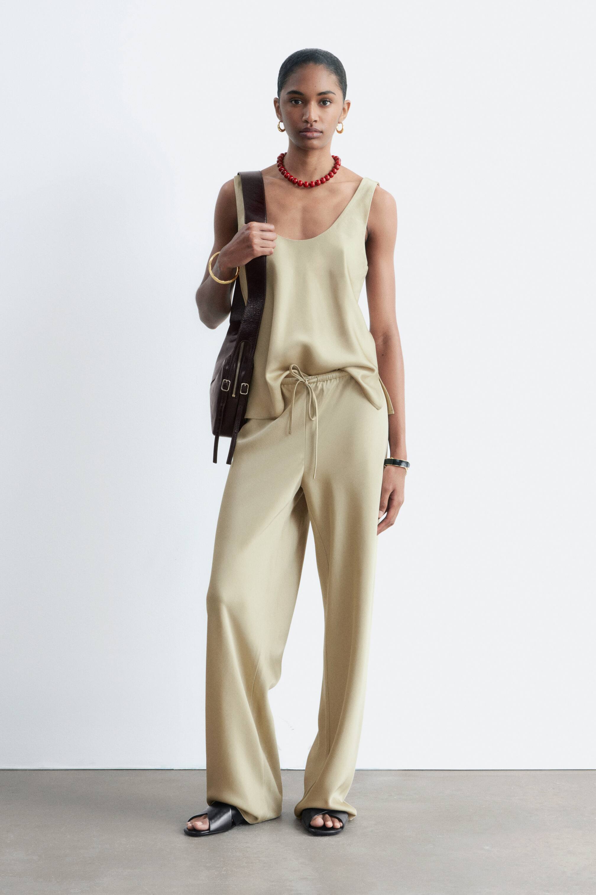 Προβολή μεγαλύτερης εικόνας: Ανοιχτό μπεζ παντελόνι με φαρδιά γραμμή, με εμφανές κορδόνι στη μέση και ρευστή πτώση, φοριέται από το μοντέλο. Το παντελόνι συνδυάζεται με ασορτί καμιζόλ και μαύρα σανδάλια.