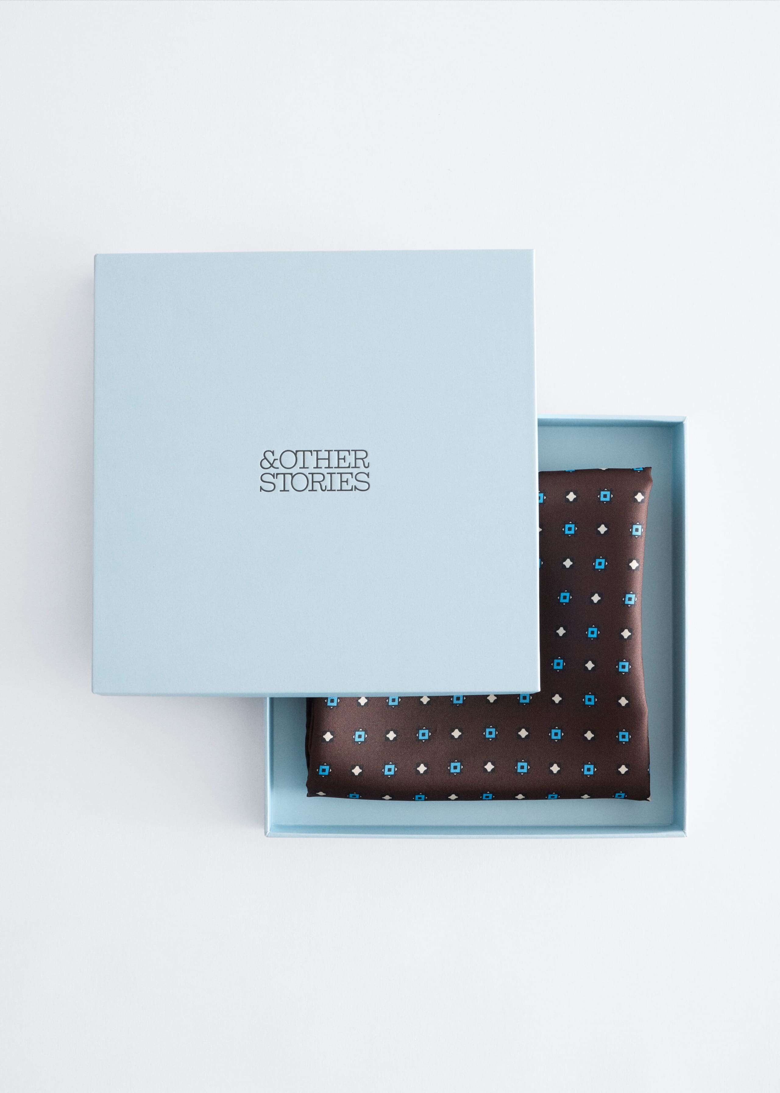 A folded silk scarf with a dark brown base and an all-over geometric pattern of small blue squares and white diamonds rests inside a light blue square gift box. The matching light blue lid, featuring the "& OTHER STORIES" logo centered on top, is partially slid to the side to reveal the folded scarf.