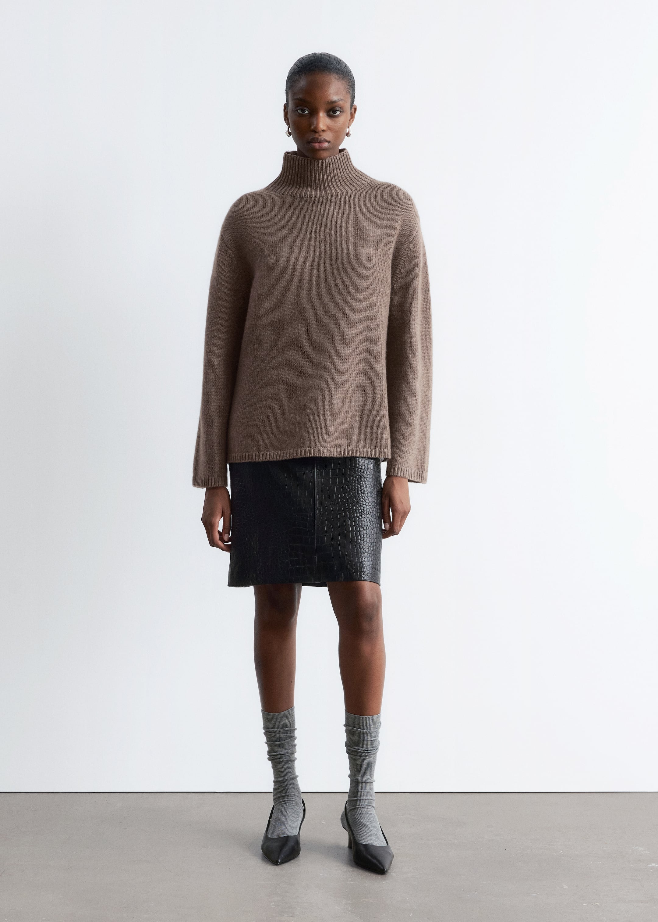 Grösseres Bild ansehen: Ein hellbrauner, locker sitzender Pullover wird präsentiert, der einen gerippten Rollkragen, lange Ärmel und einen gerippten Saum aufweist. Ein schwarzer, krokogeprägter Minirock wird unter dem Pullover getragen, der sein strukturiertes Muster und den geraden Schnitt zur Schau stellt.