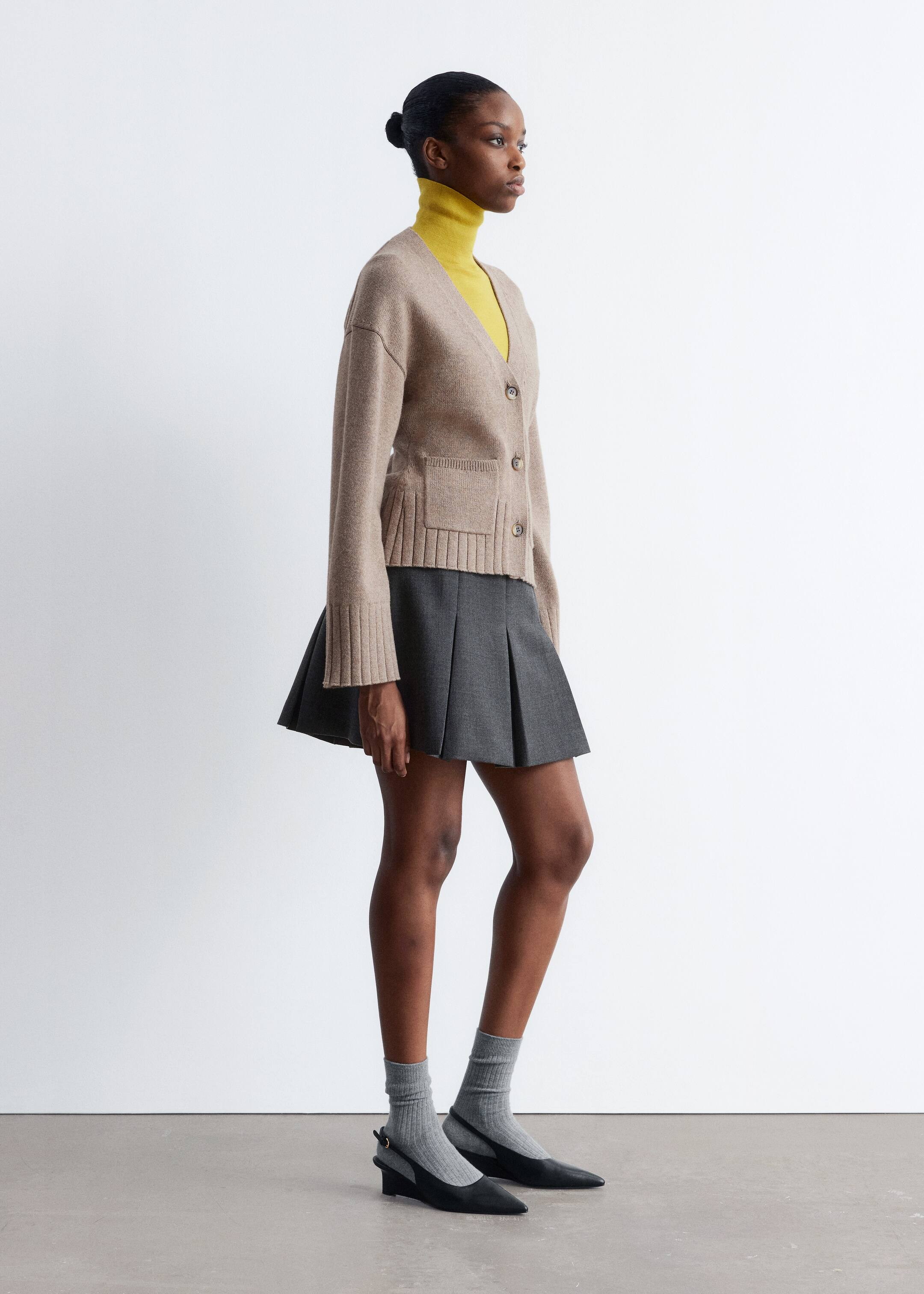 Größeres Bild anzeigen: Ein beige V-Ausschnitt-Cardigan wird in einer Seitenansicht präsentiert, mit einer Vorderseite mit drei Knöpfen, zwei aufgesetzten Taschen und gerippten Bündchen und Saum, offen über einem gelben Rollkragenpullover gestylt, um seine hüftlange Silhouette zu zeigen.