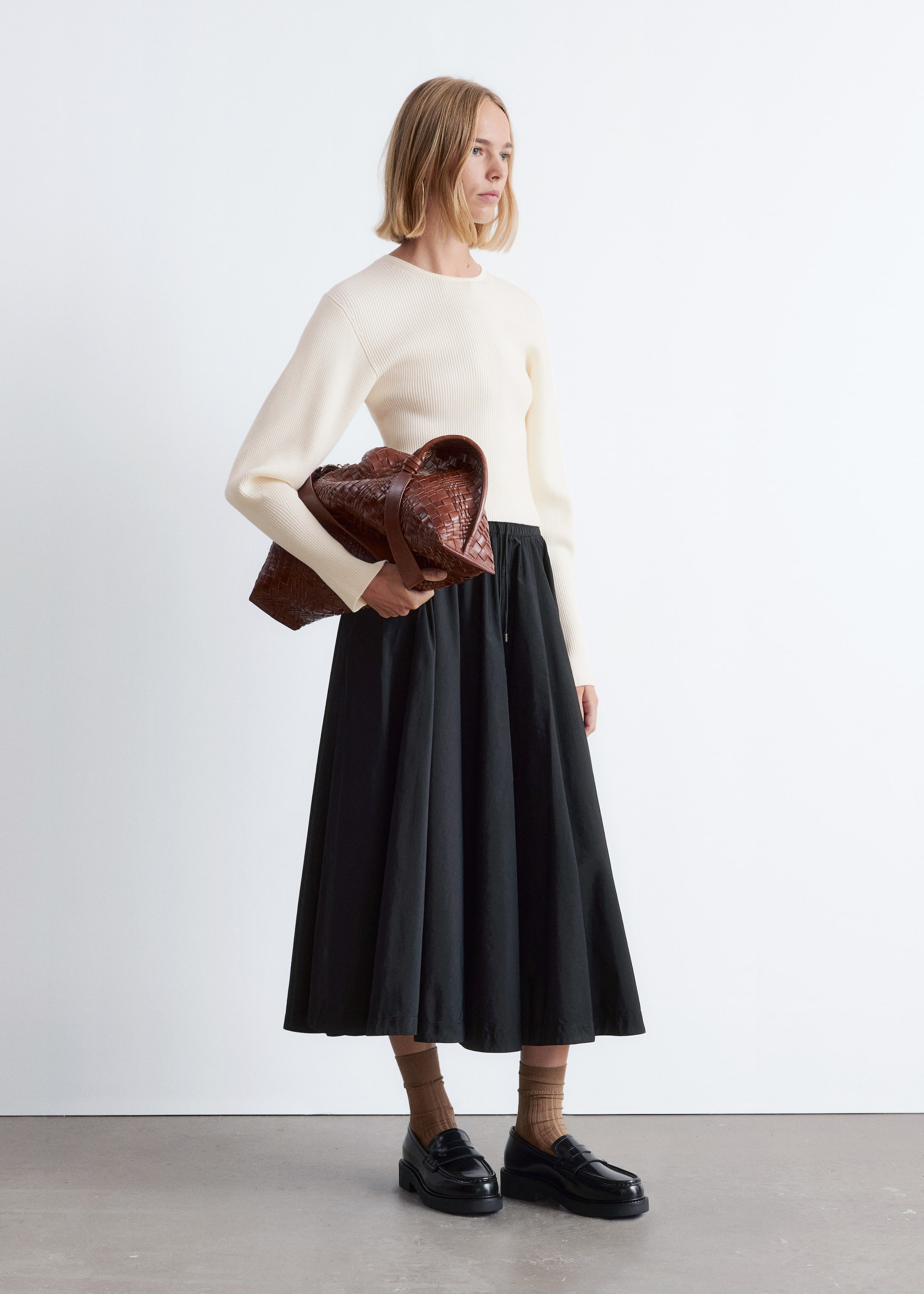 Grösseres Bild ansehen: Ein schwarzer Midirock präsentiert sich mit einer vollen, ausgestellten Silhouette und einem sichtbaren Kordelzug an der Taille. Er wird kombiniert mit einem cremeweißen gerippten Strickpullover, einer braunen geflochtenen Ledertasche, braunen gerippten Socken und schwarzen klobigen Loafern.
