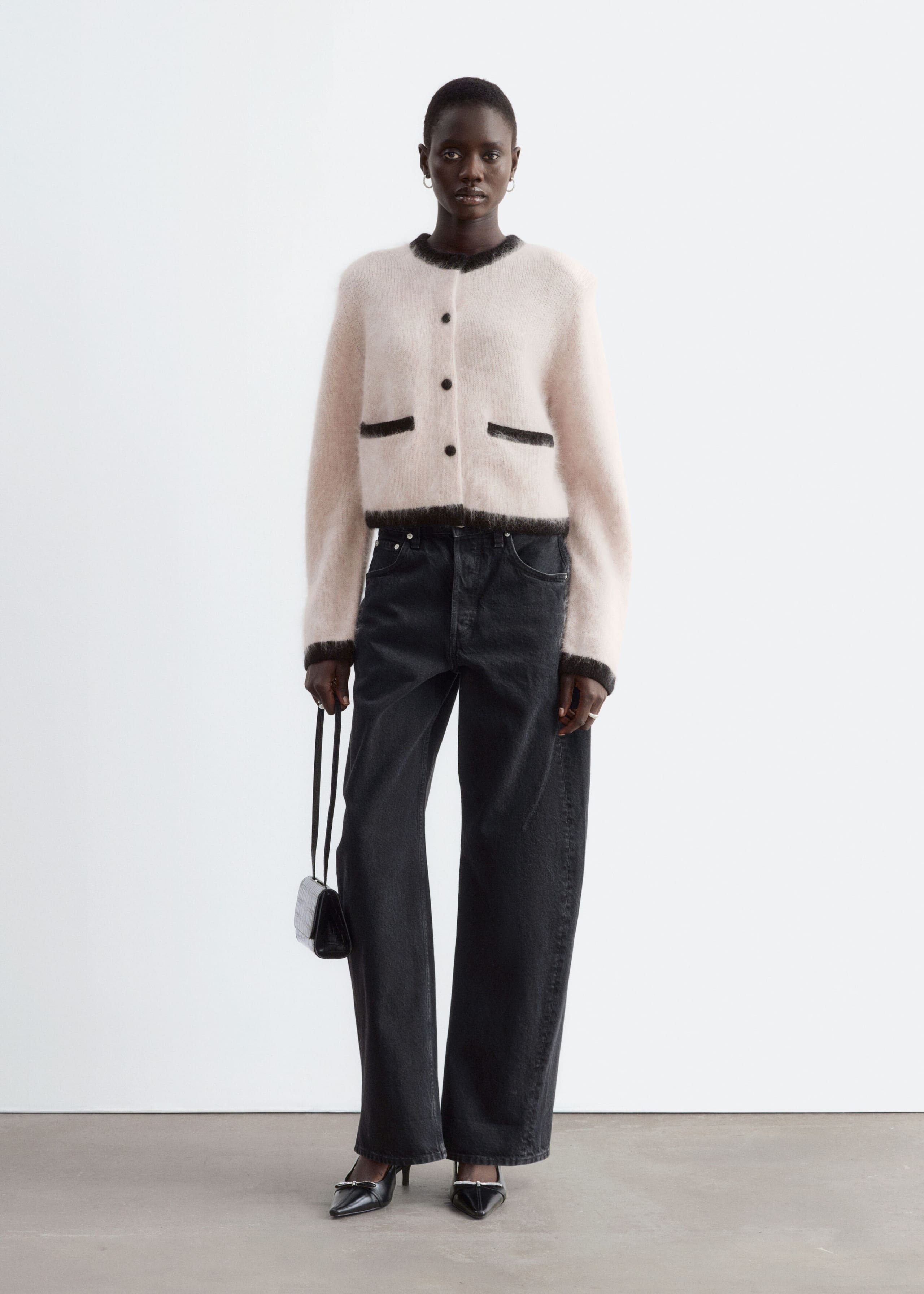 A fuzzy, pale pink cardigan featuring structured shoulders, contrasting black ribbed trim, and dark front buttons is worn by the model. The cardigan is styled with wide-leg black jeans, and the model is holding a small black handbag.