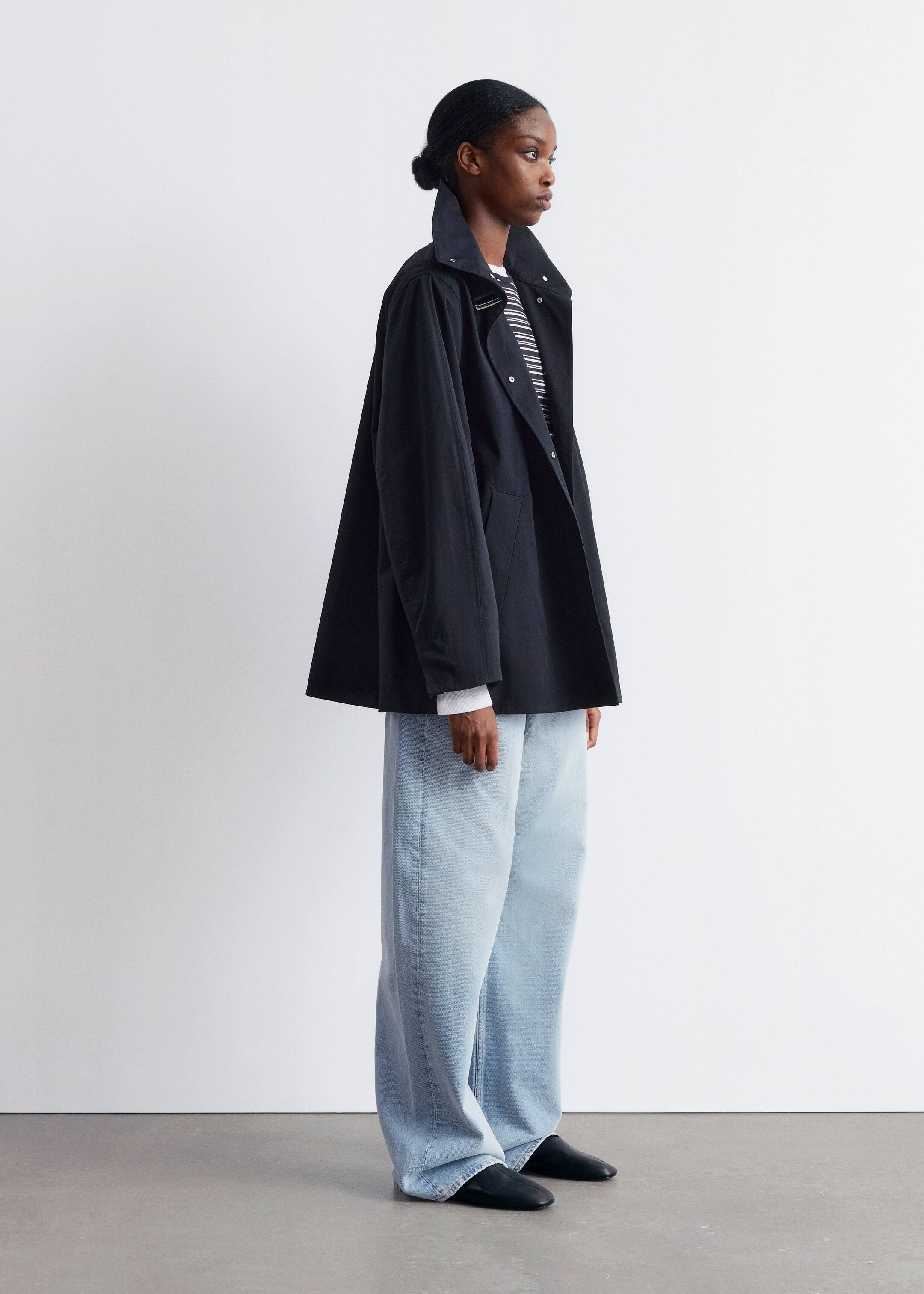 Grösseres Bild ansehen: Eine schwarze, übergroße Jacke wird im Seitenprofil getragen, mit einem hohen Stehkragen mit sichtbaren Druckknöpfen und einer breiten A-Linien-Silhouette, die über einem schwarz-weiß gestreiften Oberteil und weißen Hemdmanschetten geschichtet ist, wobei hellblaue, weit geschnittene Jeans den entspannten Look vervollständigen.