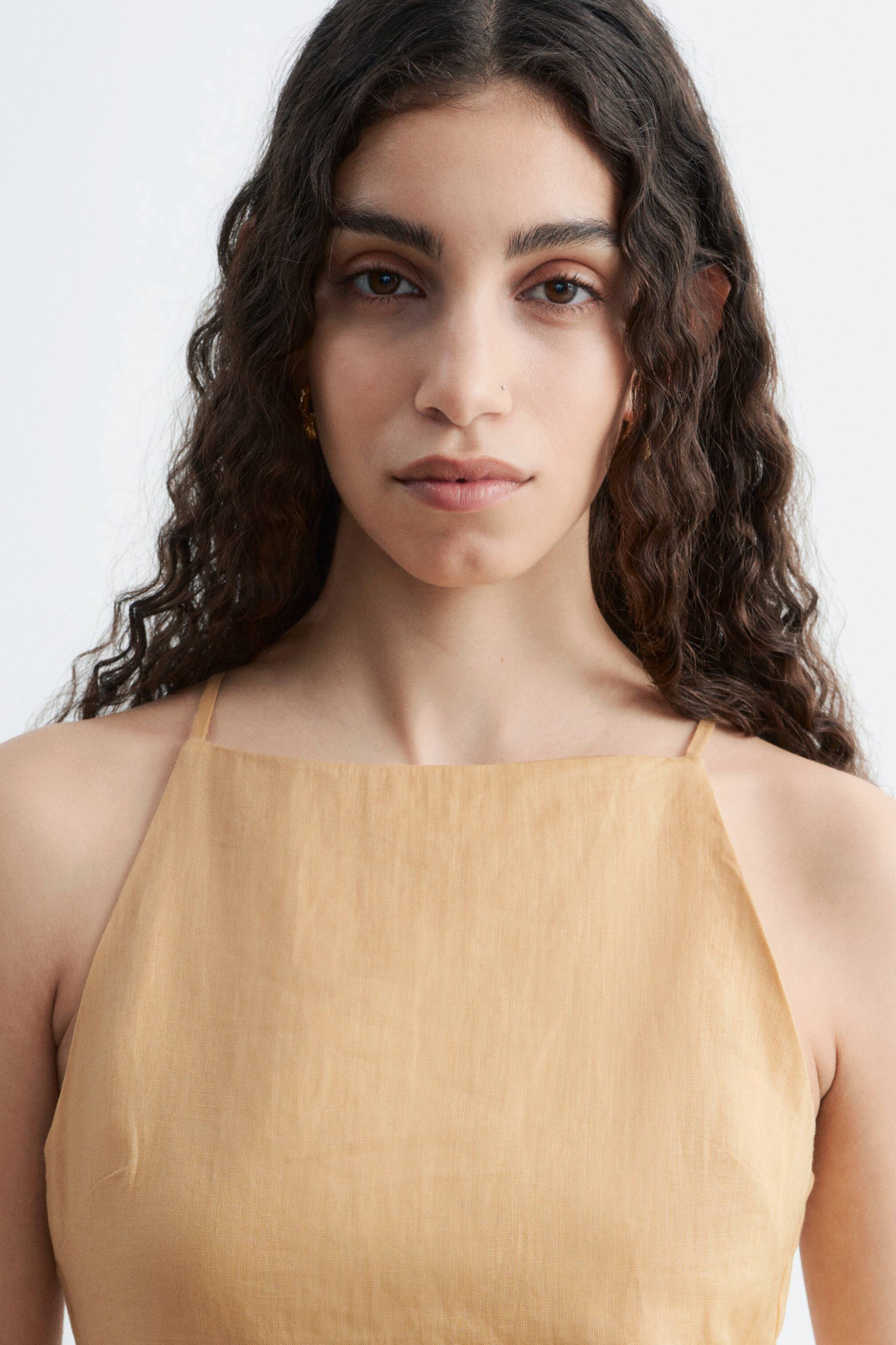 Visualizza immagine più grande: Un abito di lino color camoscio è indossato dalla modella. Presenta un collo alto e sottili spalline.