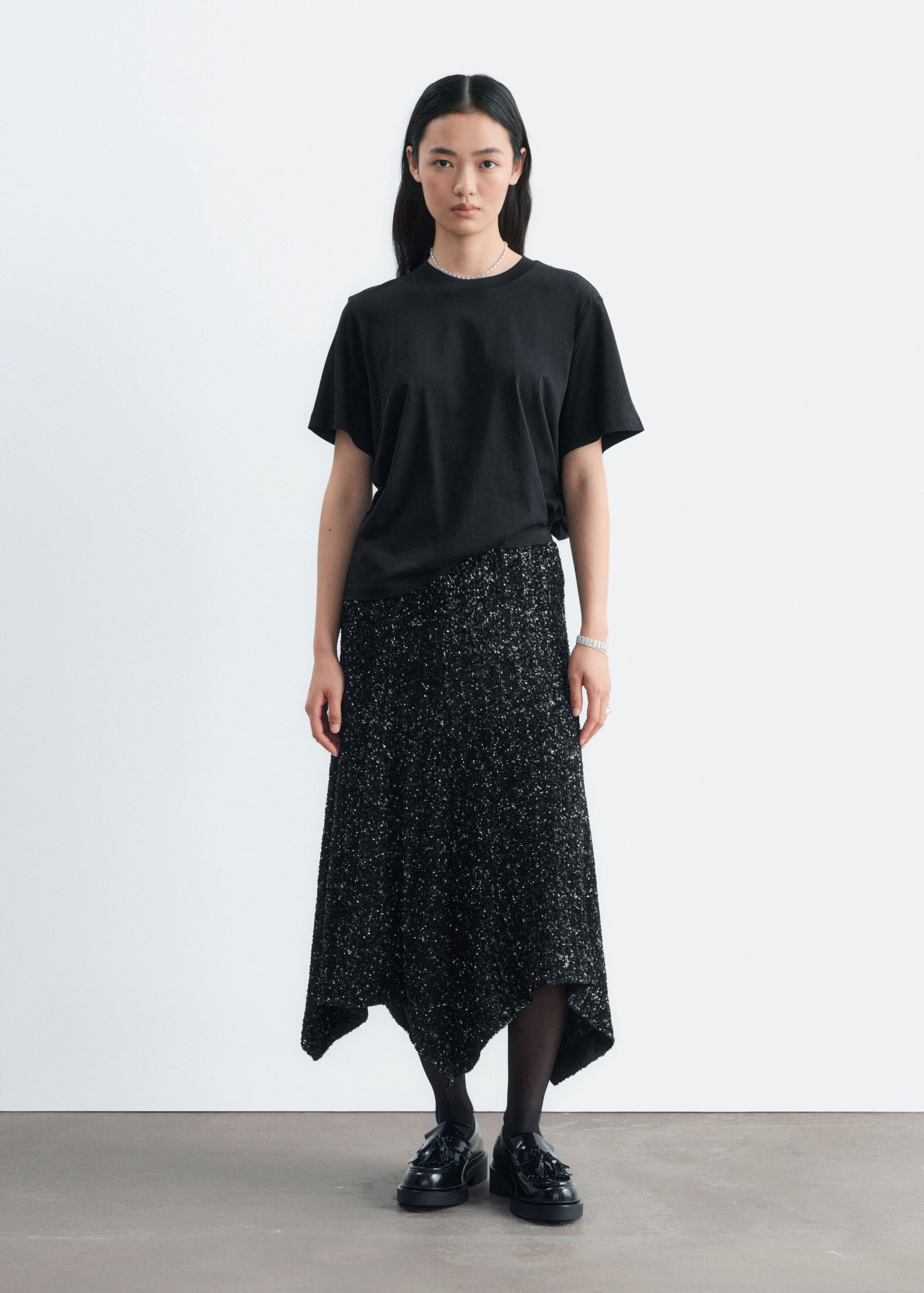 The model is wearing a black, knee-length sequined mesh skirt with an asymmetrical hem. It is styled with a tucked-in black T-shirt, black tights, and black leather loafers.