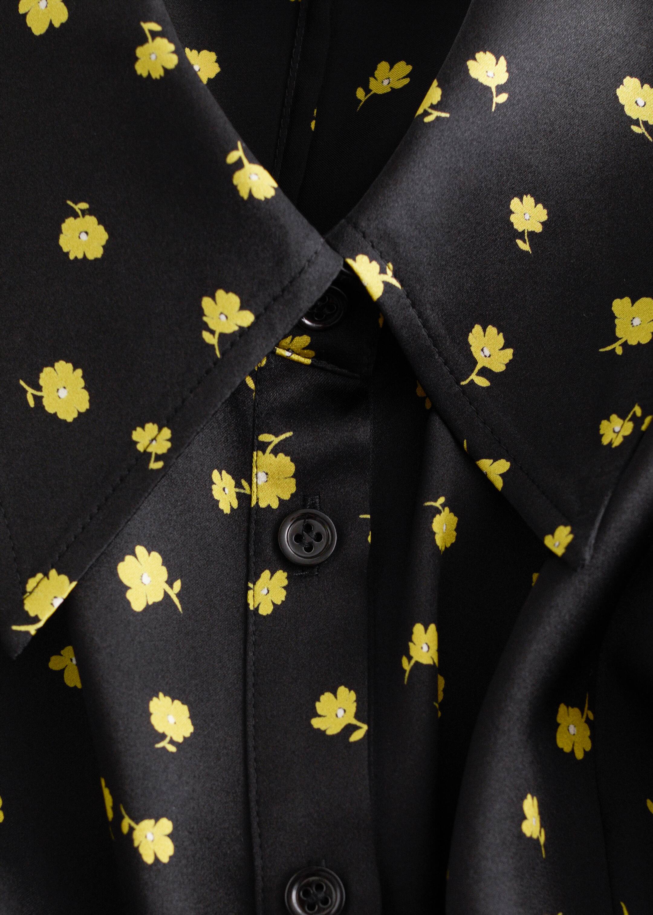 Ver imagem maior: Uma peça de vestuário preta, semelhante a cetim, adornada com um estampado floral amarelo disperso, está a ser mostrada numa vista aproximada e angulada. Apresenta uma gola pontiaguda e uma carcela de botões à frente, presa com botões escuros de quatro furos.
