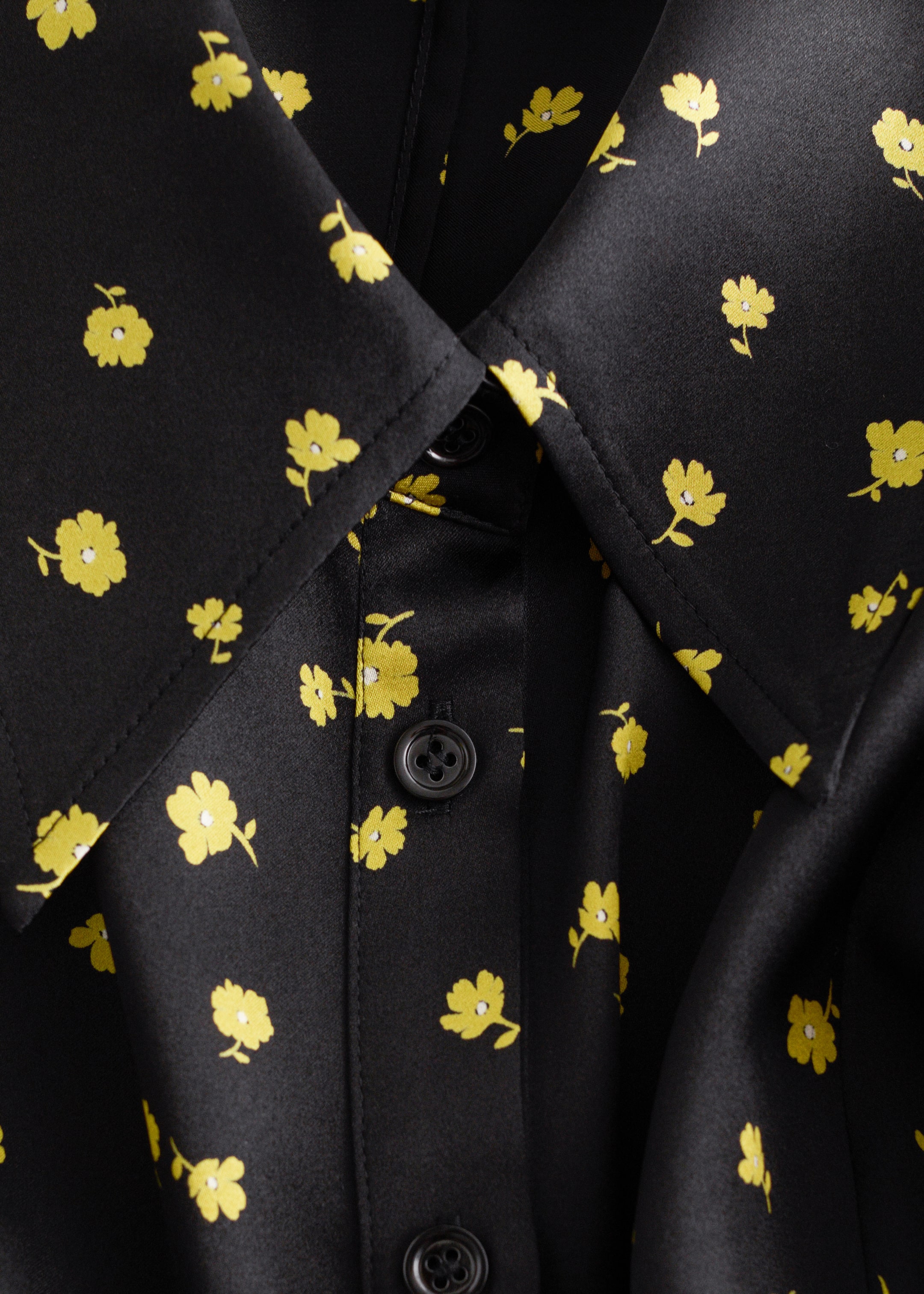 View larger image: A black, satin-like garment, adorned with a scattered yellow floral print, is being shown in a close-up, angled view. It is featuring a pointed collar and a front button placket fastened with dark four-hole buttons.