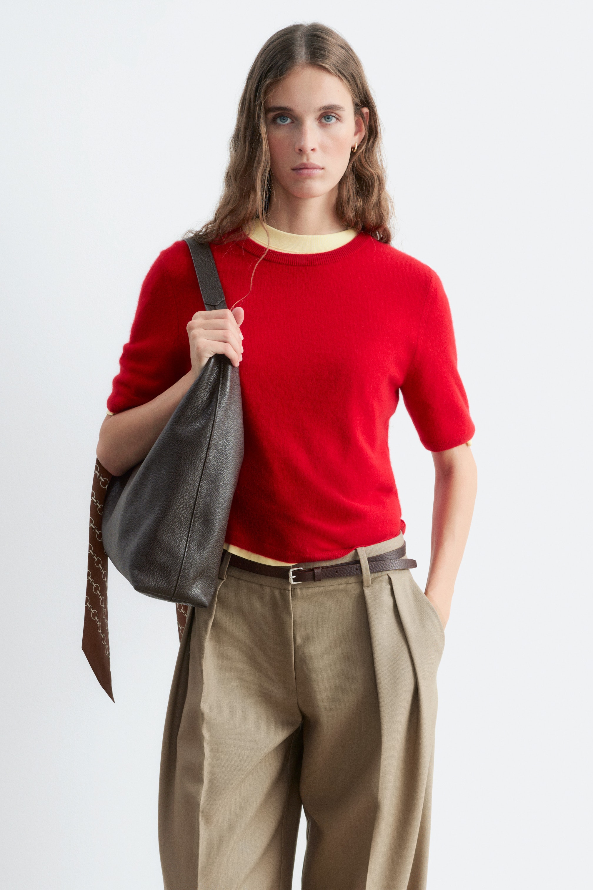 Ver imagem maior: A modelo está a usar uma camisola de caxemira vermelha de manga curta, sobreposta a uma camisola de gola redonda amarelo-claro. A peça de malha apresenta gola redonda canelada e está combinada com calças chino caqui com pregas e uma mala de ombro em pele castanha.