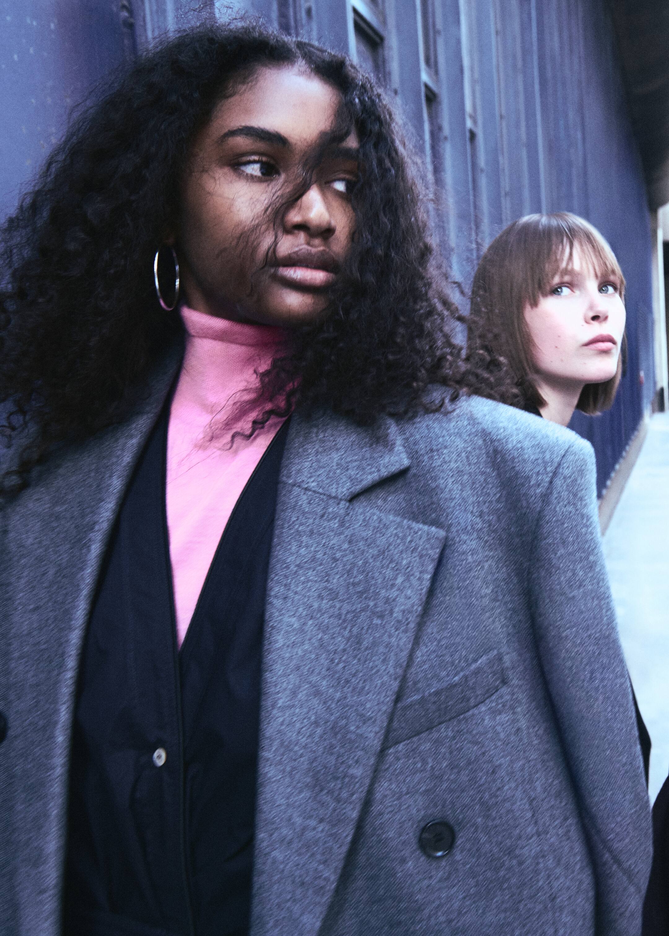 Größeres Bild anzeigen: Ein grauer Blazer mit entspannter Passform wird getragen, der breite Kerbrevers und einen sichtbaren dunklen Knopfverschluss an der Vorderseite aufweist. Er ist über einem leuchtend pinken Rollkragenpullover und einem schwarzen V-Ausschnitt-Kleidungsstück mit einem weißen Knopf geschichtet.