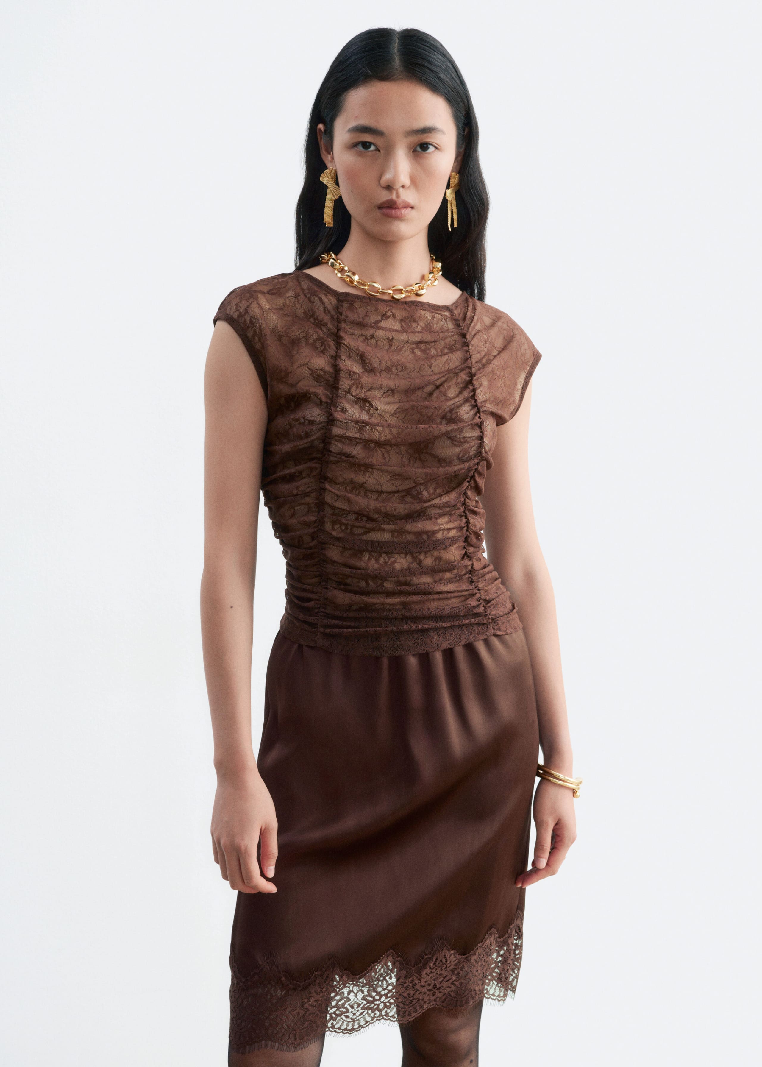 A brown lace top featuring vertical ruching through the body, a boat neckline, and cap sleeves is worn by the model. The top is styled with a matching brown satin skirt with a lace-trimmed hem.