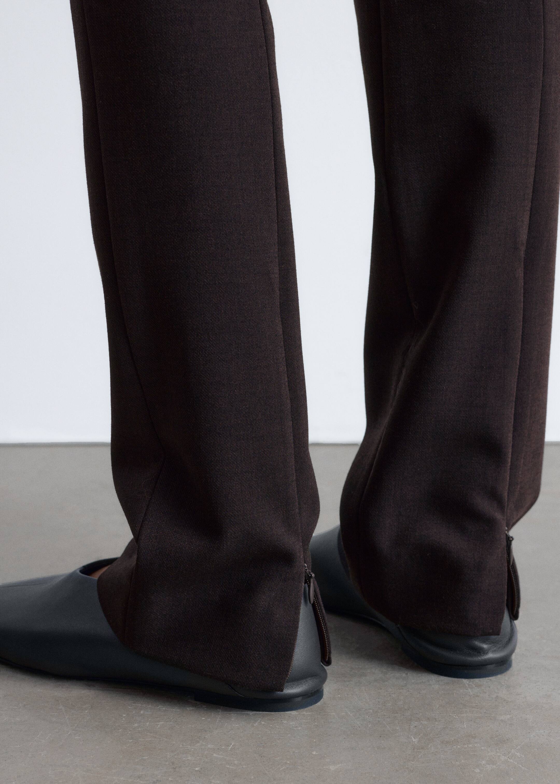 Agrandir l'image: Un pantalon marron foncé est présenté sous un angle bas, avec une jambe droite et une texture tissée subtile. Les ourlets tombent sur des chaussures plates et sombres, et chaque cheville est dotée d'une fermeture éclair visible à l'arrière.