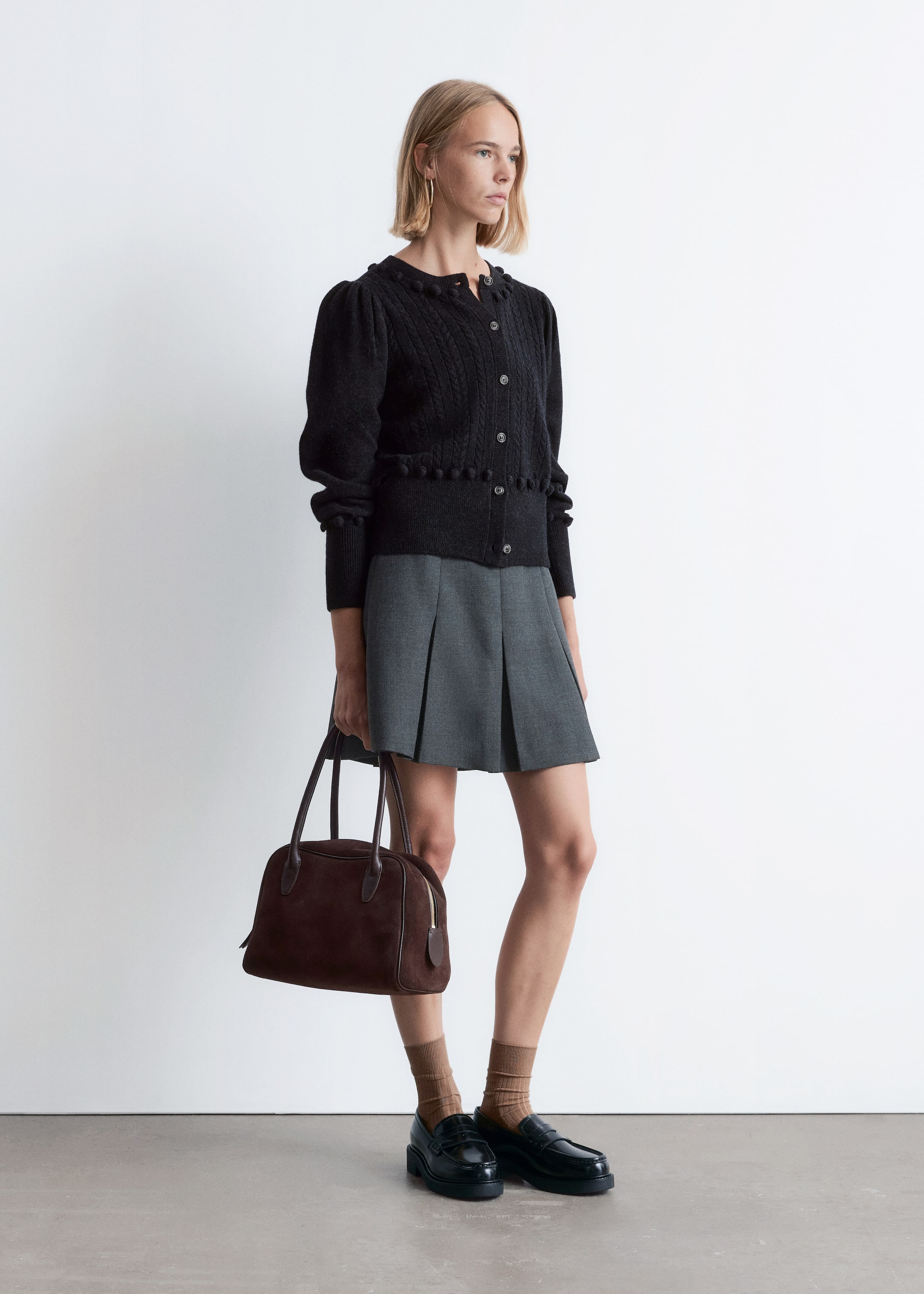 View larger image: A black cable-knit cardigan is being worn over a grey pleated skirt, facing slightly to the right. The cardigan is showcasing a round neckline, front button closure, long slightly puffed sleeves with ribbed cuffs, and a decorative pom-pom trim along its bottom hem.