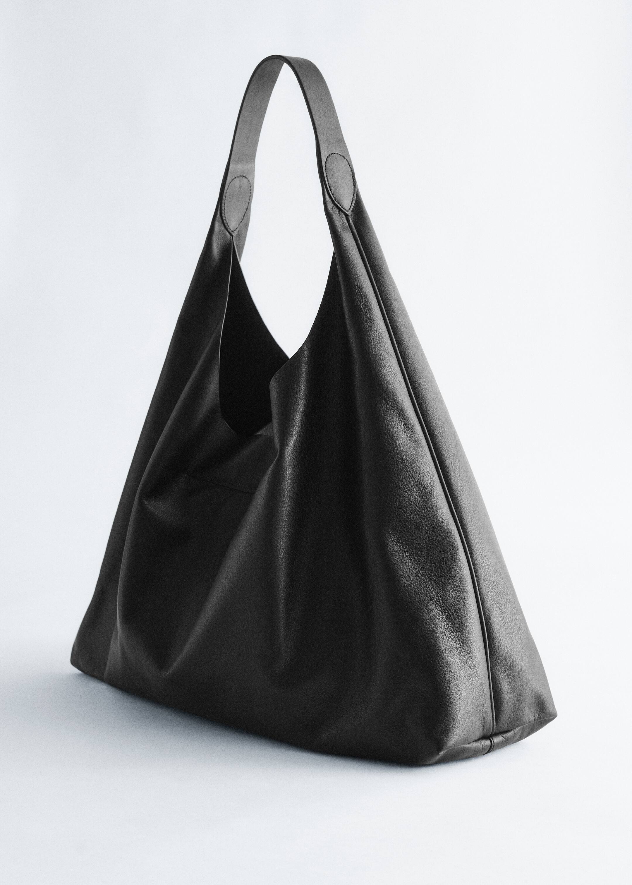 Agrandir l'image: Un sac cabas en cuir noir présente une texture douce et froissée, avec une large poignée unique s'élevant du haut. Le sac affiche une grande ouverture souple et une base structurée.