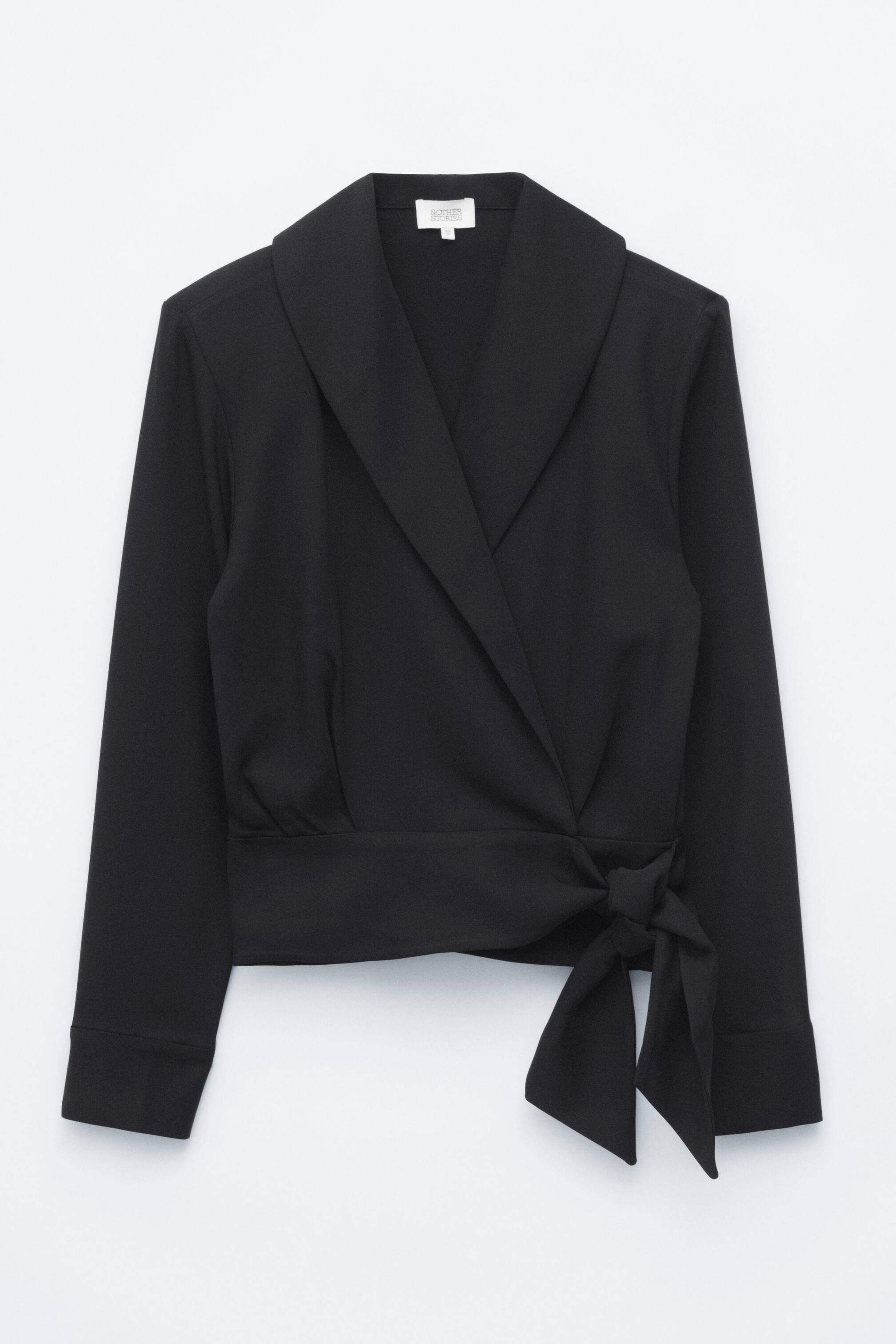 Grotere afbeelding bekijken: Een zwarte wikkelblouse met lange mouwen wordt van voren getoond, met een brede sjaalkraag en een omwikkeld silhouet. De blouse wordt aan de zijkant van de taille vastgemaakt met een strik van dezelfde stof, waardoor zachte plooien boven de tailleband ontstaan.