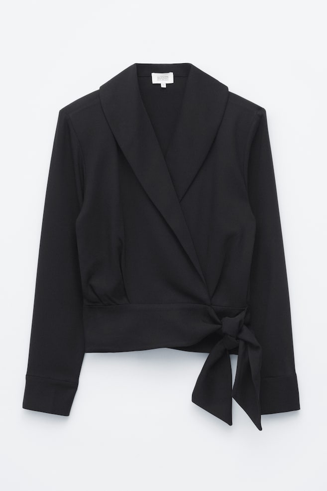 Een zwarte wikkelblouse met lange mouwen wordt van voren getoond, met een brede sjaalkraag en een omwikkeld silhouet. De blouse wordt aan de zijkant van de taille vastgemaakt met een strik van dezelfde stof, waardoor zachte plooien boven de tailleband ontstaan. - 1