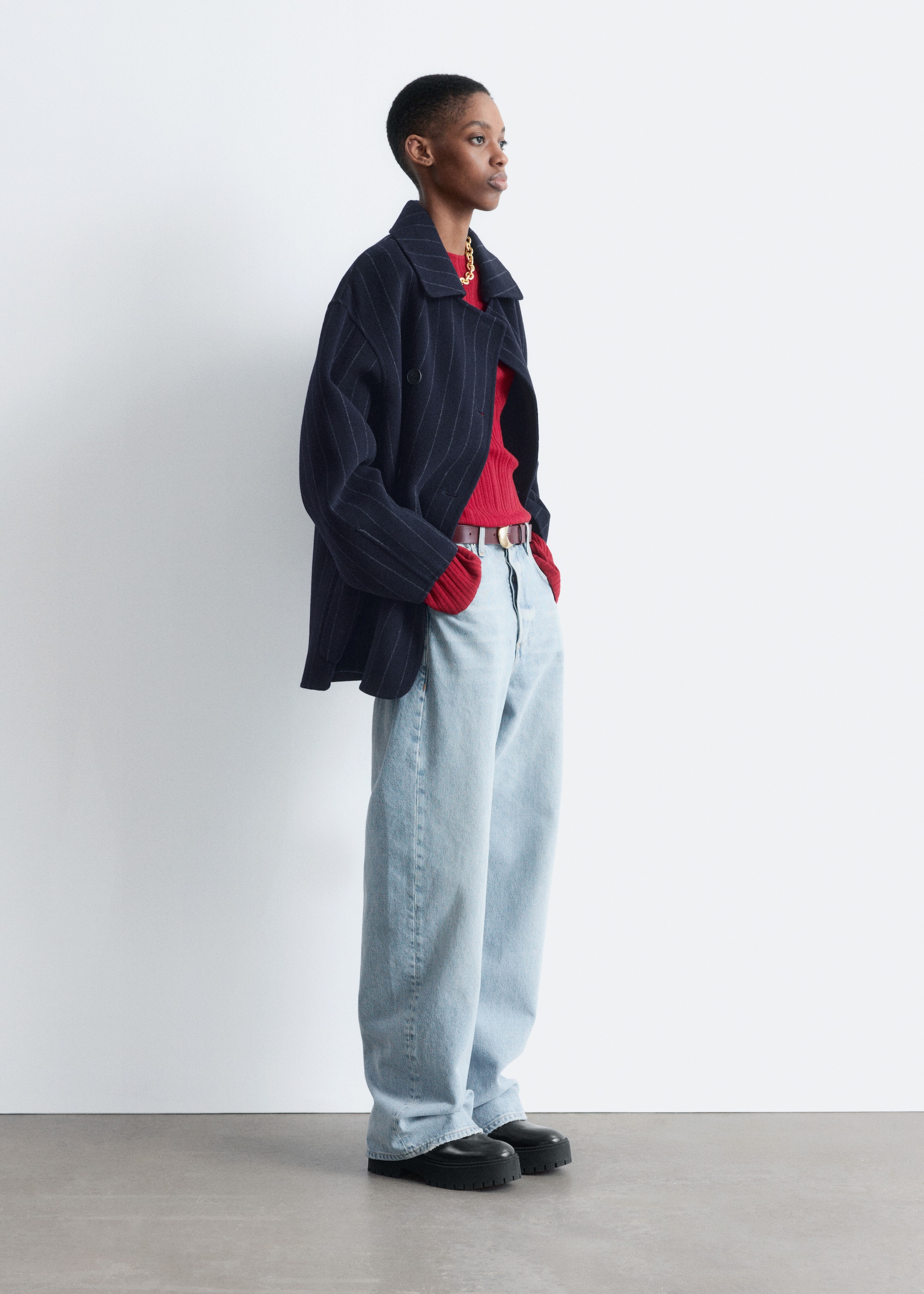 Visualizza immagine più grande: Una persona indossa una giacca doppiopetto blu navy a righe sottili con revers classici, sovrapposta a un top rosso a coste, jeans a gamba larga chiari, una cintura marrone con fibbia dorata e mocassini neri con suola spessa, rivolta verso destra.
