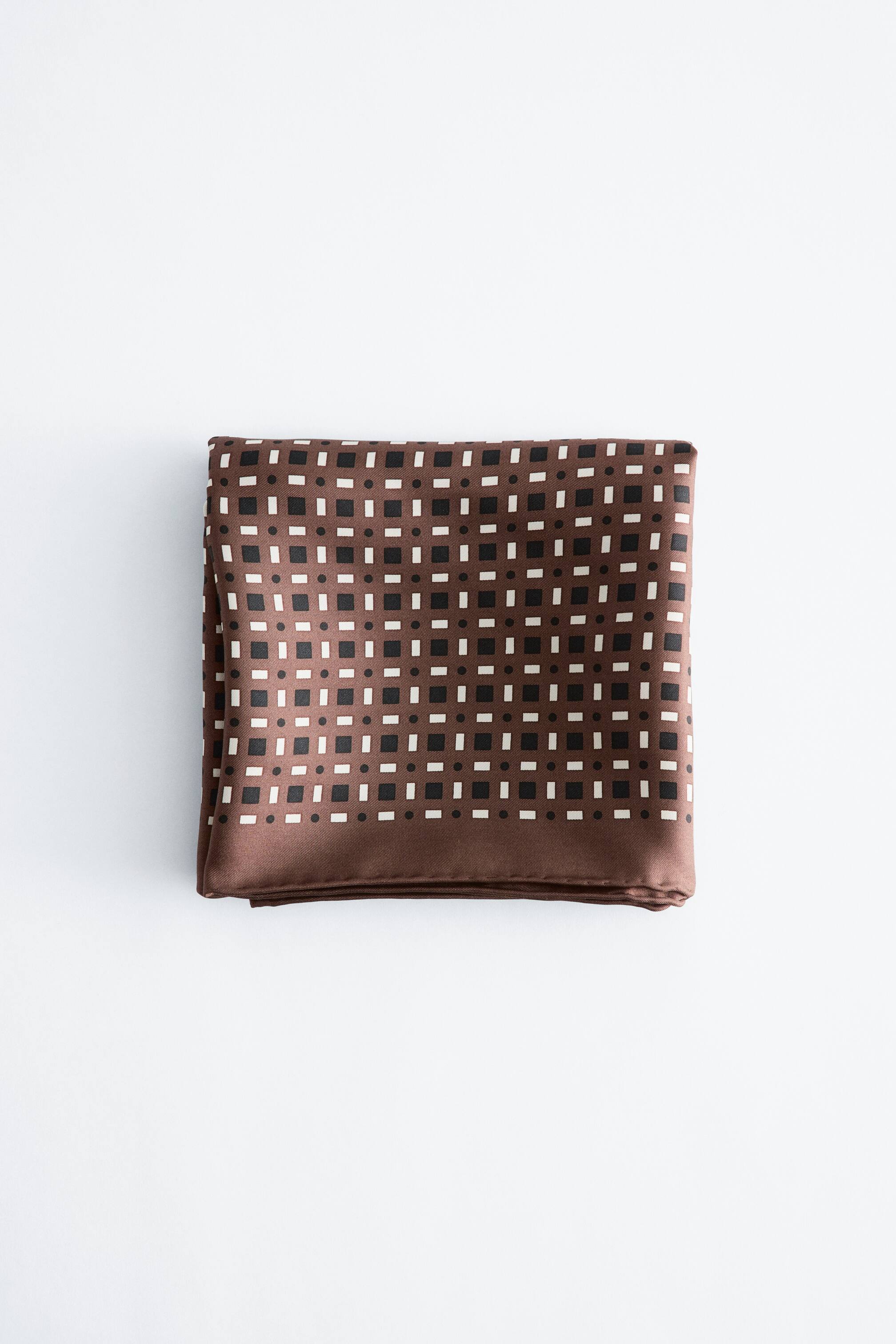 View larger image: A brown scarf is folded into a rectangular shape, showcasing a repeating geometric pattern. The design features small black squares, white rectangles, and white dots.