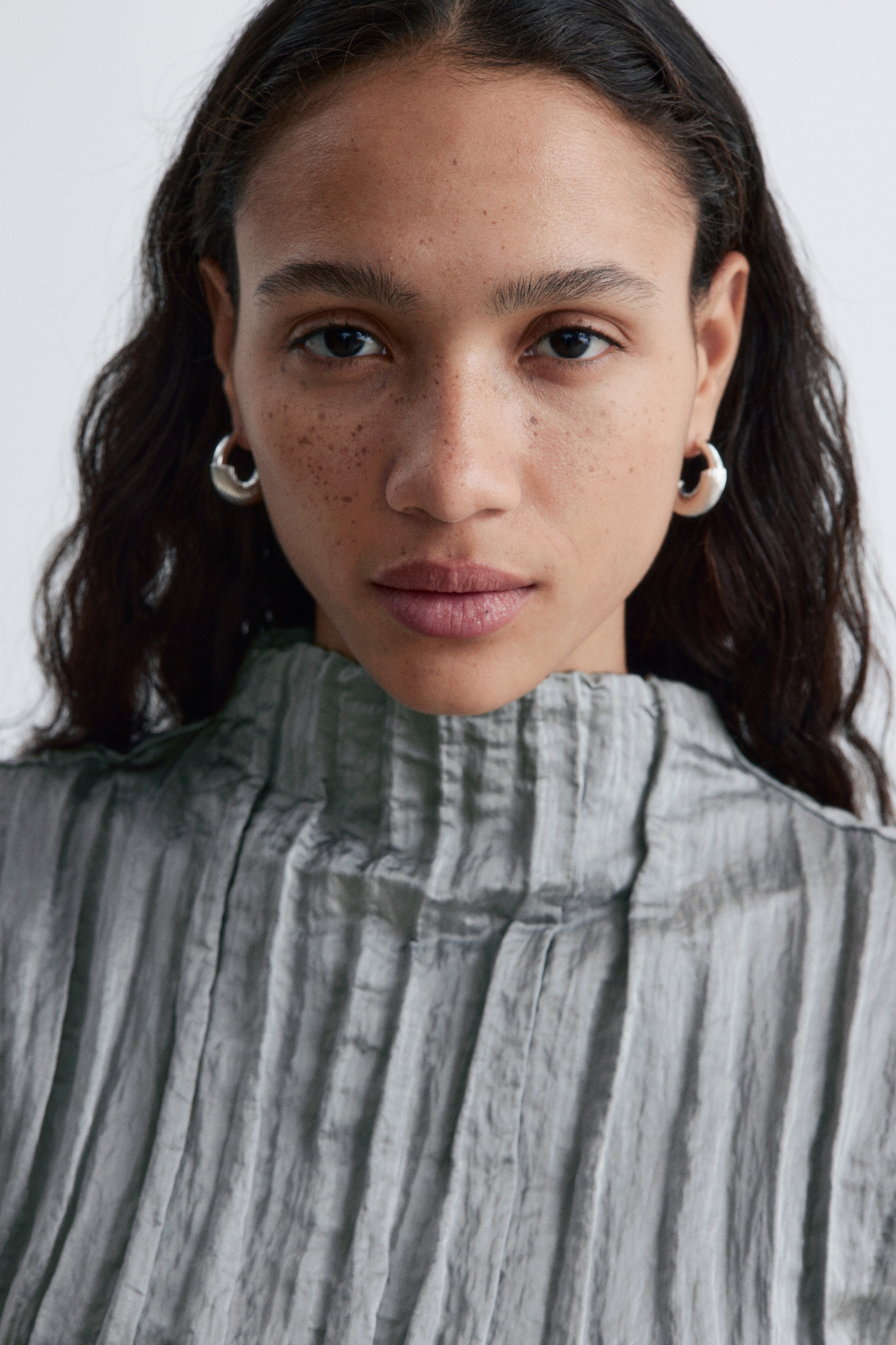 Grotere afbeelding bekijken: Een grijze, hooggesloten top met verticale plooien en een gestructureerde, gekreukelde stof, gepresenteerd vanuit een vooraanzicht. Dikke, zilverkleurige, ronde hoepeloorbellen sieren beide oorlellen en tonen hun volledige vorm vanuit een vooraanzicht.