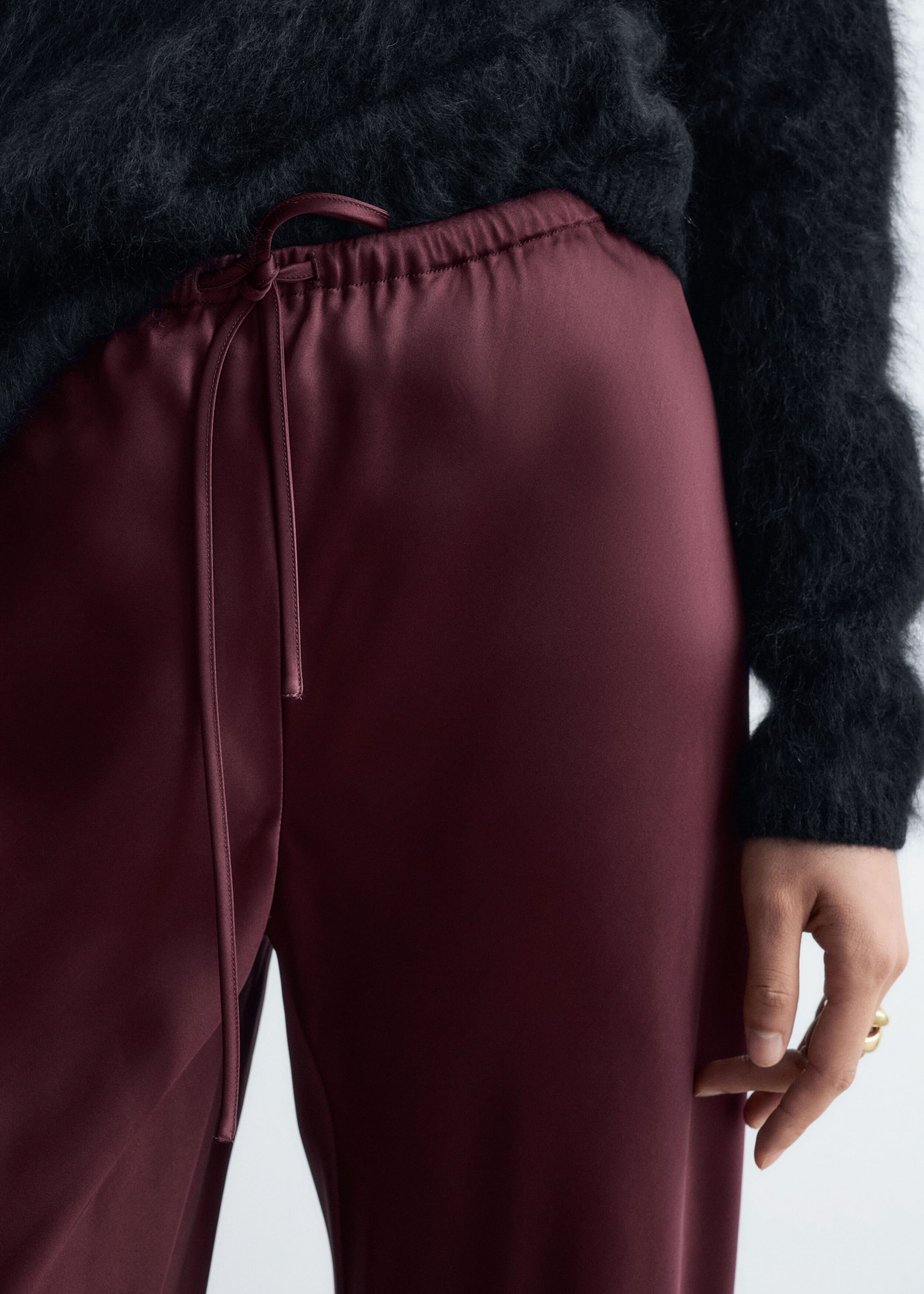 The model is wearing burgundy satin trousers featuring an elastic waistband with a matching drawstring tie. The trousers are styled with a black fuzzy knit sweater tucked into the waist.