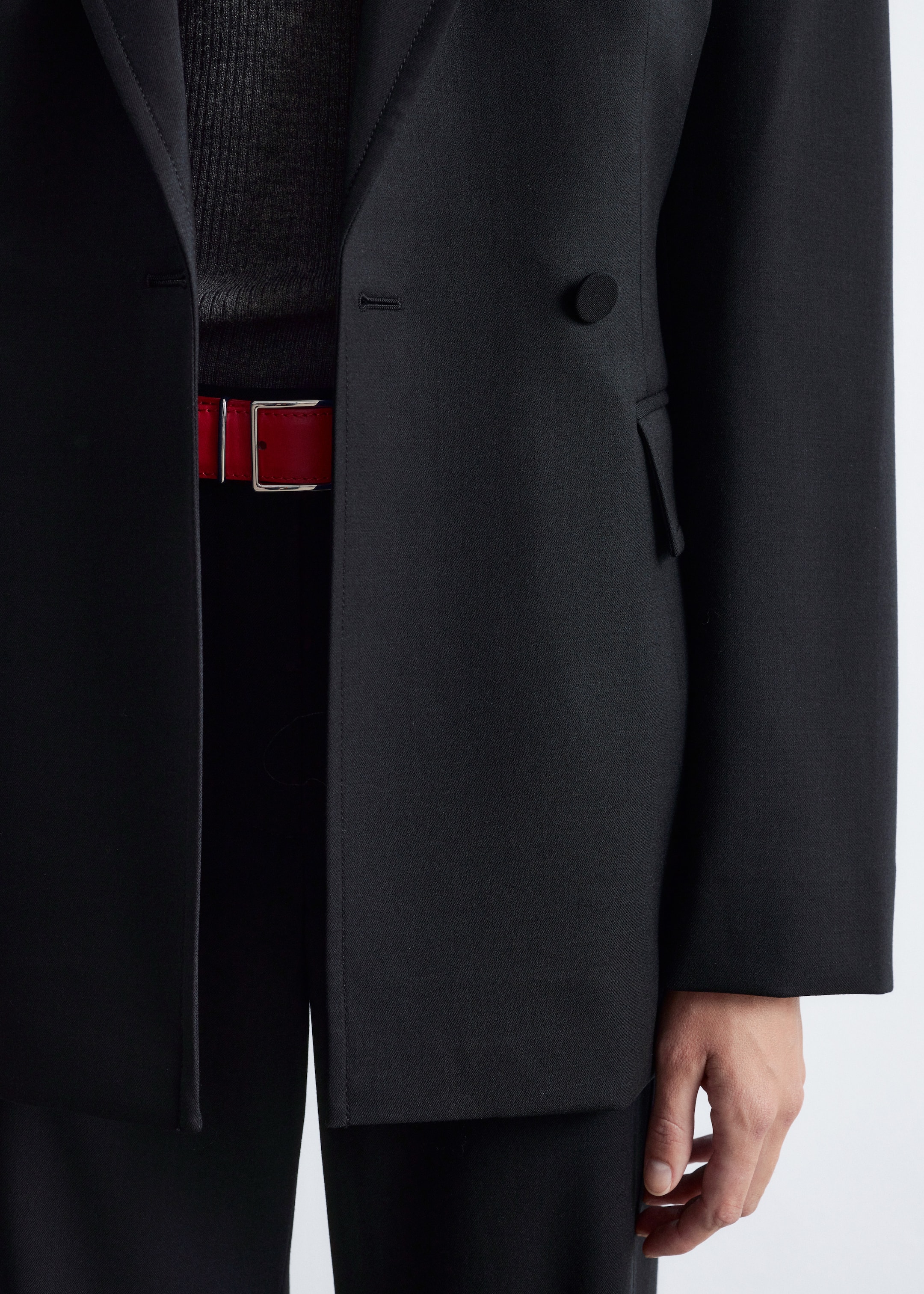 Agrandir l'image: Un blazer noir à double boutonnage est présenté de face, avec un bouton noir sur le côté droit, une boutonnière visible sur le côté gauche et une poche à rabat. Il est porté ouvert, révélant un haut côtelé gris et une ceinture rouge avec une boucle argentée.