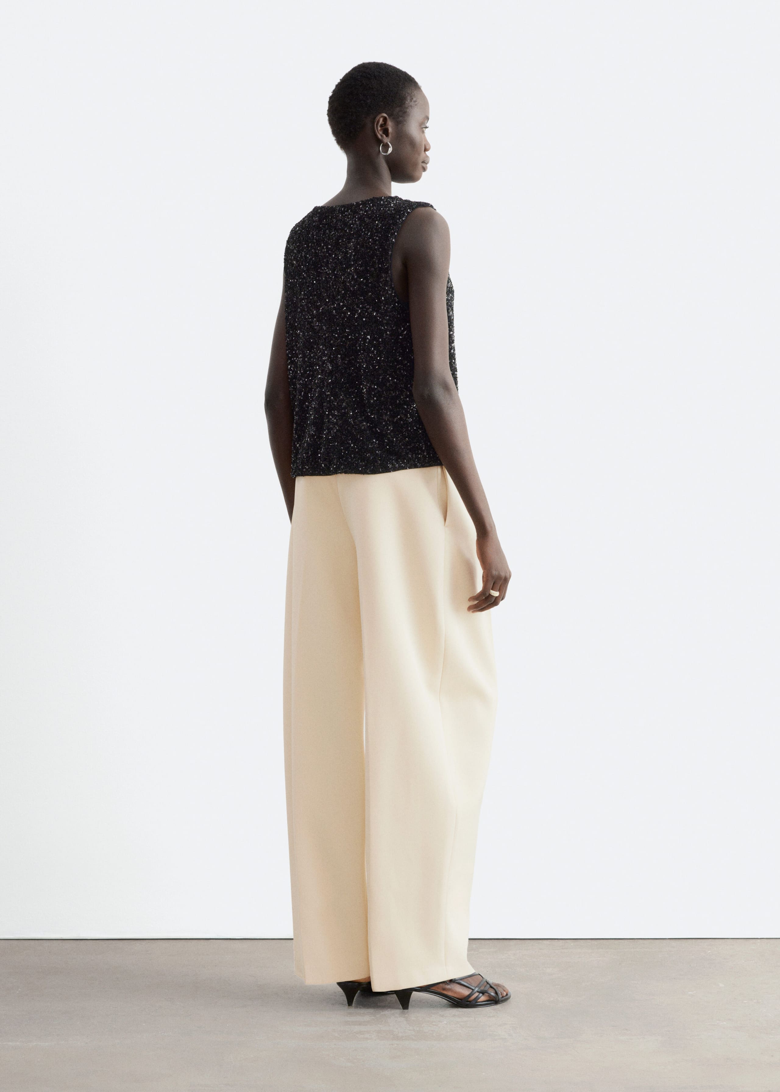A black, sleeveless sequinned top is being worn by the model, shown from a rear three-quarter angle. The shimmering top is styled with cream-coloured, wide-leg trousers.