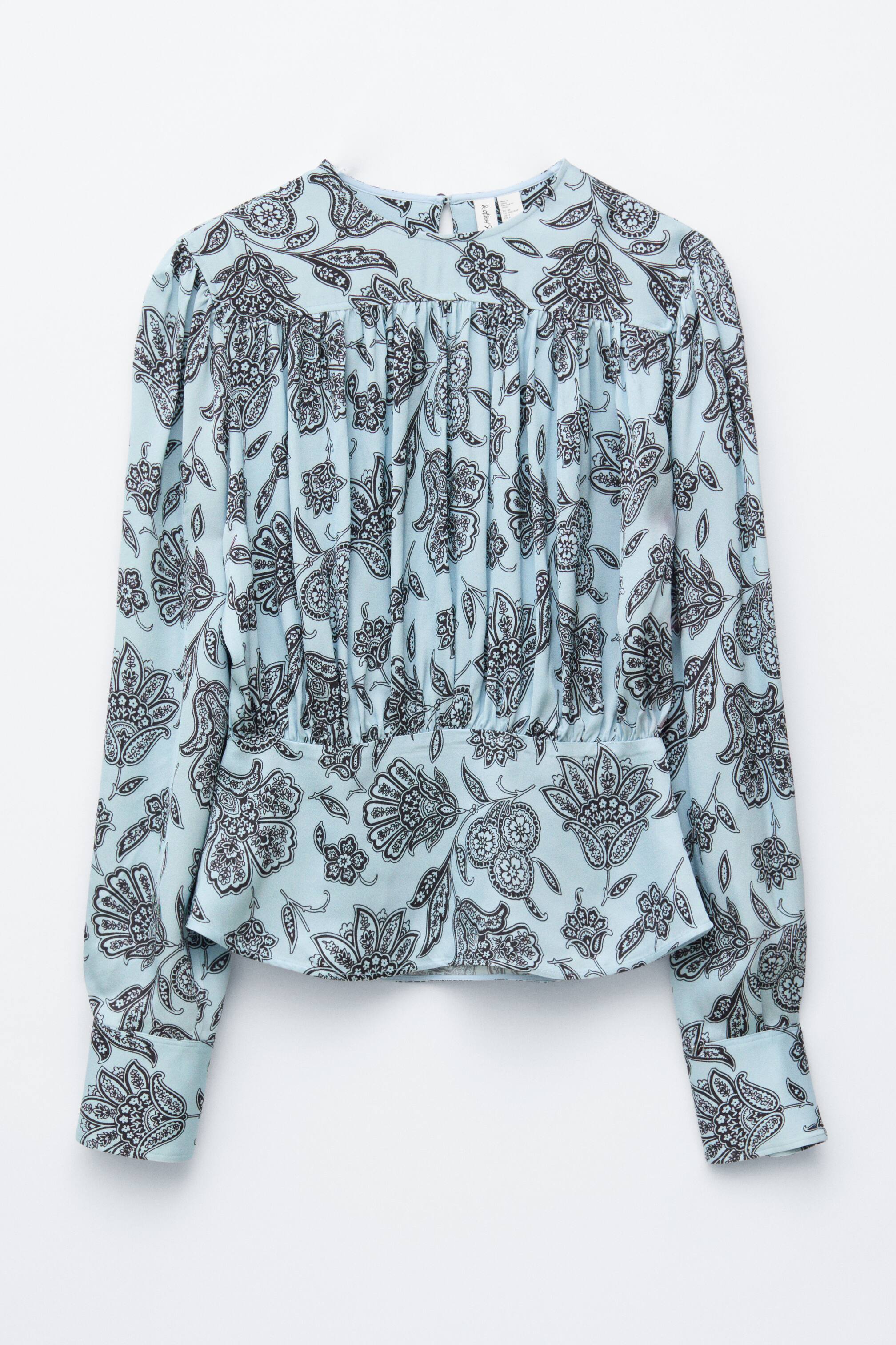 Agrandir l'image: Une blouse bleu clair est présentée de FACE, arborant un motif floral et cachemire répétitif en gris foncé et noir. Elle présente un col rond haut, un plissage étendu sur le haut de la poitrine, et une taille cintrée créant un effet blouson. Des manches longues avec des poignets boutonnés complètent le design.