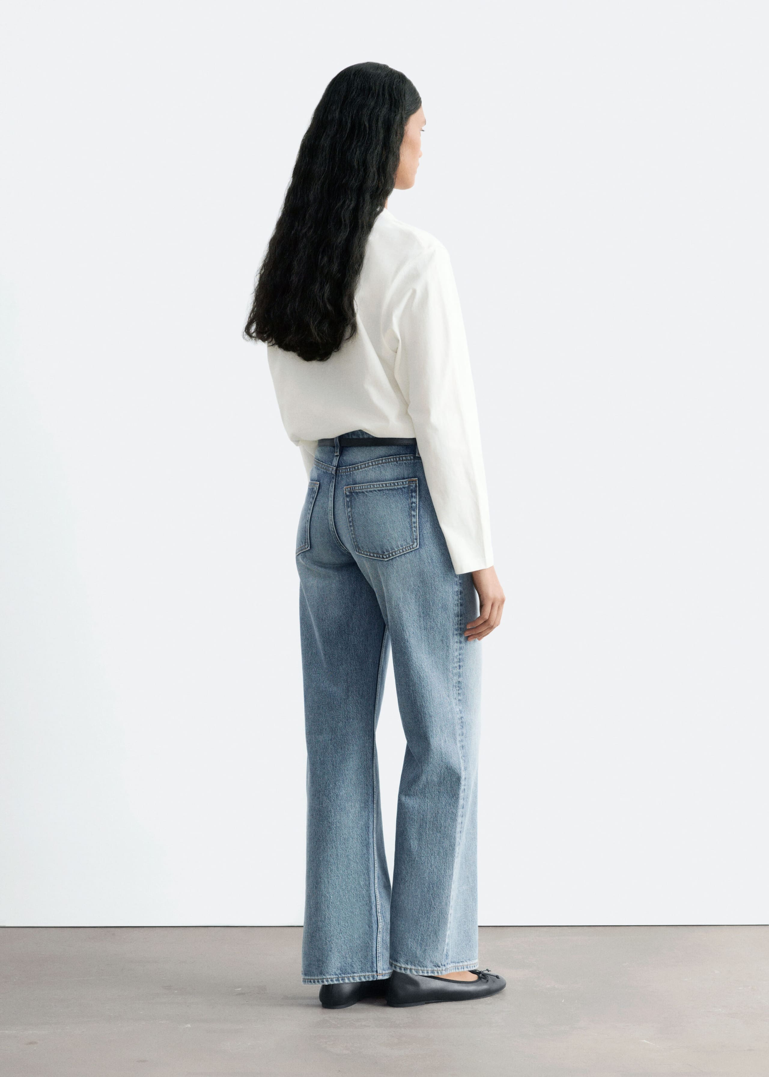 The model is wearing medium-wash, straight-leg jeans, shown from a three-quarter rear angle to highlight the mid-rise waist, regular fit and classic back pockets. The jeans are styled with a tucked-in white long-sleeved shirt and black flat shoes.
