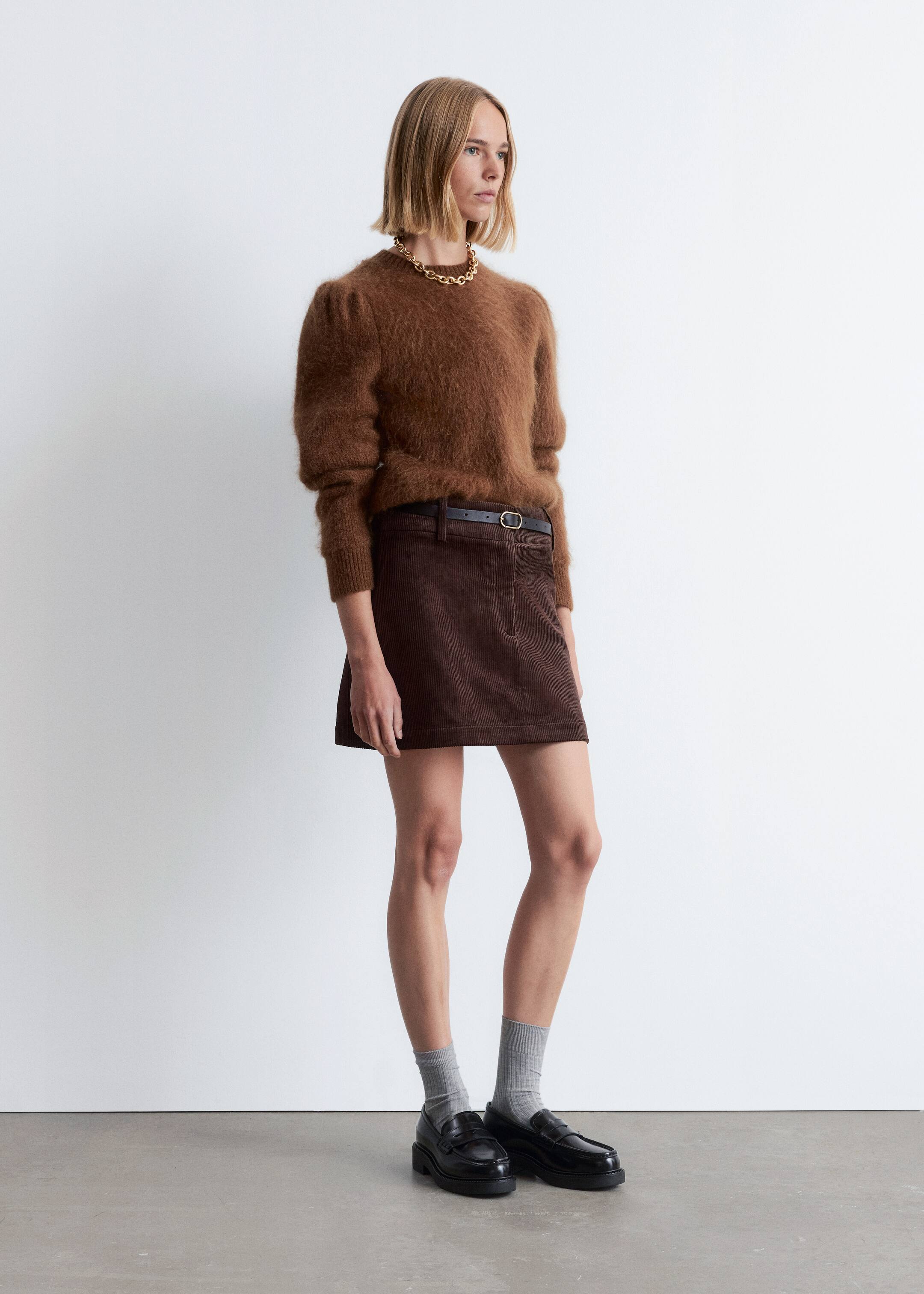 View larger image: A brown fuzzy sweater with voluminous sleeves and a banded hem is being shown from a side angle. It is being paired with a brown corduroy A-line mini skirt, which is styled with a thin black belt featuring a gold buckle.