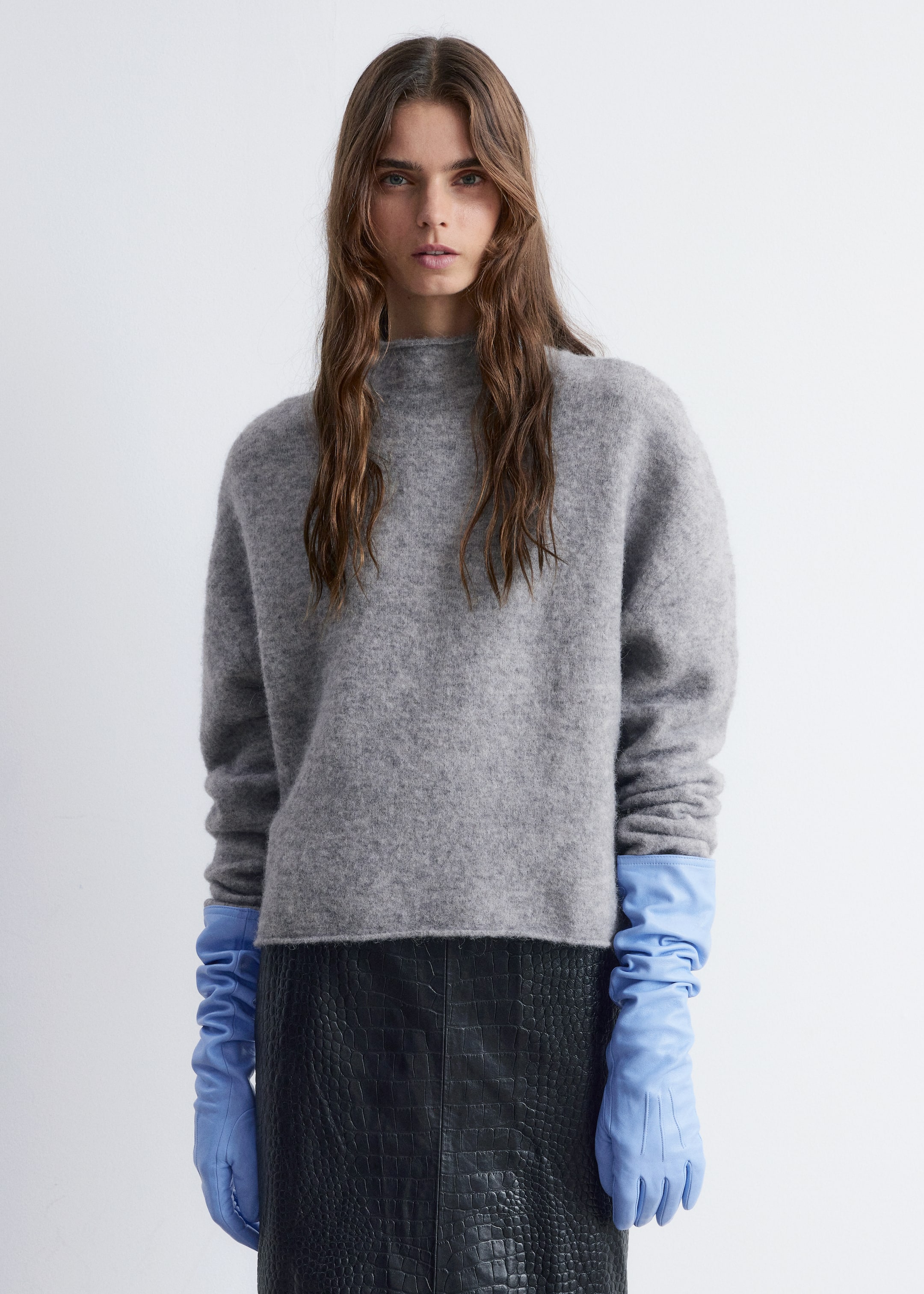 Visa större bild: En grå mock-neck tröja som bärs framåt, med en luddig textur, avslappnade långa ärmar och en rak fåll. Ljusblå läderhandskar sträcker sig ut från under tröjans ärmar, och en svart kjol med krokodilpräglat mönster syns under dess fåll.