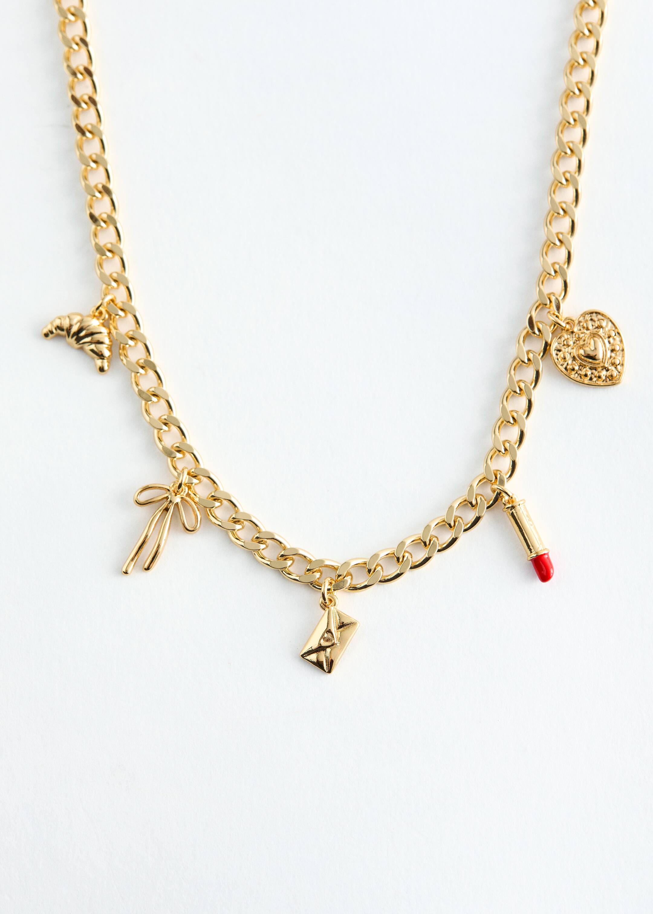 Agrandir l'image: Un collier en chaîne gourmette en laiton poli est posé à plat, affichant son DEVANT, avec cinq petits pendentifs qui pendent le long de sa longueur : un croissant, un nœud, une lettre, un rouge à lèvres avec une pointe rouge, et un cœur texturé.