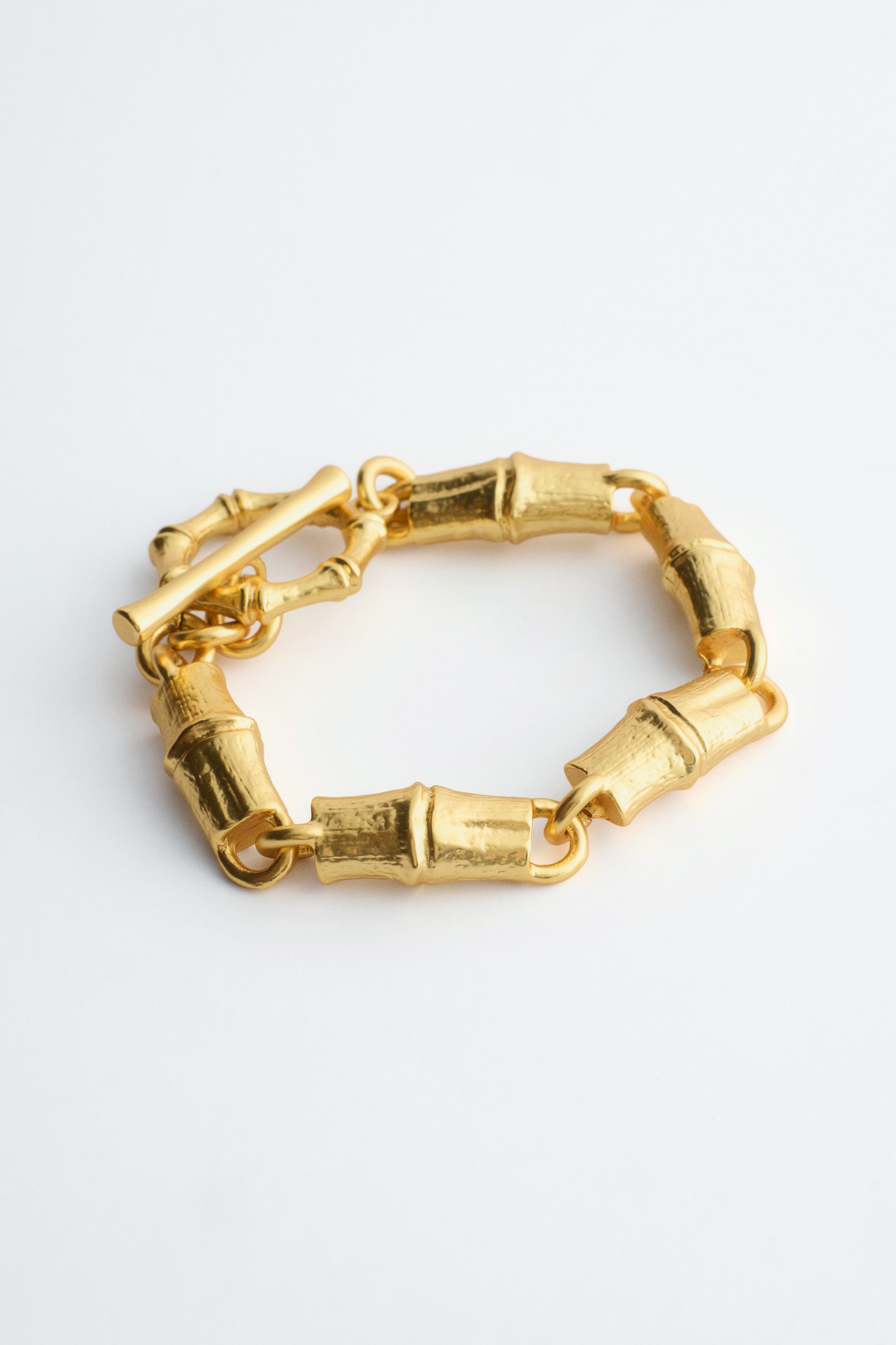 Visualizza l'immagine ingrandita: Un bracciale robusto color oro è adagiato piatto, formando una forma ovale. Presenta maglie scolpite e testurizzate che ricordano segmenti di bambù e mette in evidenza una chiusura a barra nella parte anteriore.