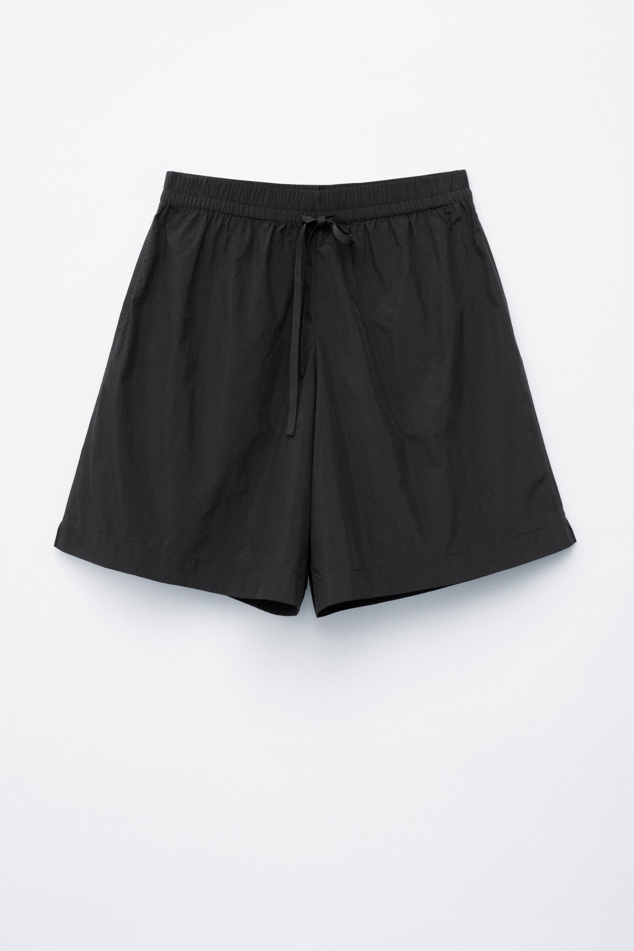View larger image: Black shorts are shown from the front, featuring a relaxed, knee-length silhouette and an elasticated waistband with a drawstring tied at the centre.