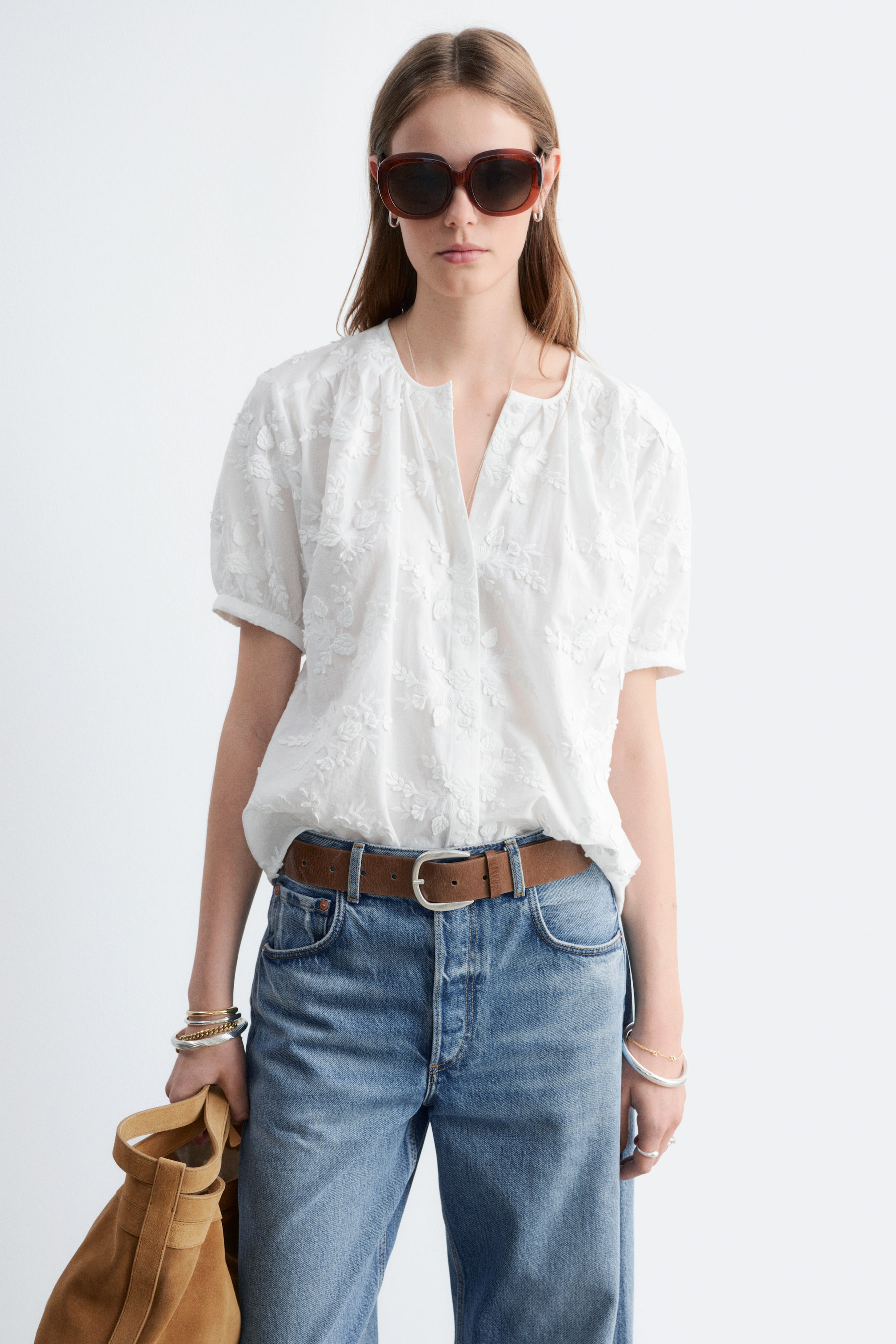 Se større billede: En hvid broderet bluse præsenterer en V-udskæring med en delvis knaplukning og puffede korte ærmer. Den fremviser indviklet blomsterbroderi over stoffet og er delvist stukket ind i blå jeans.