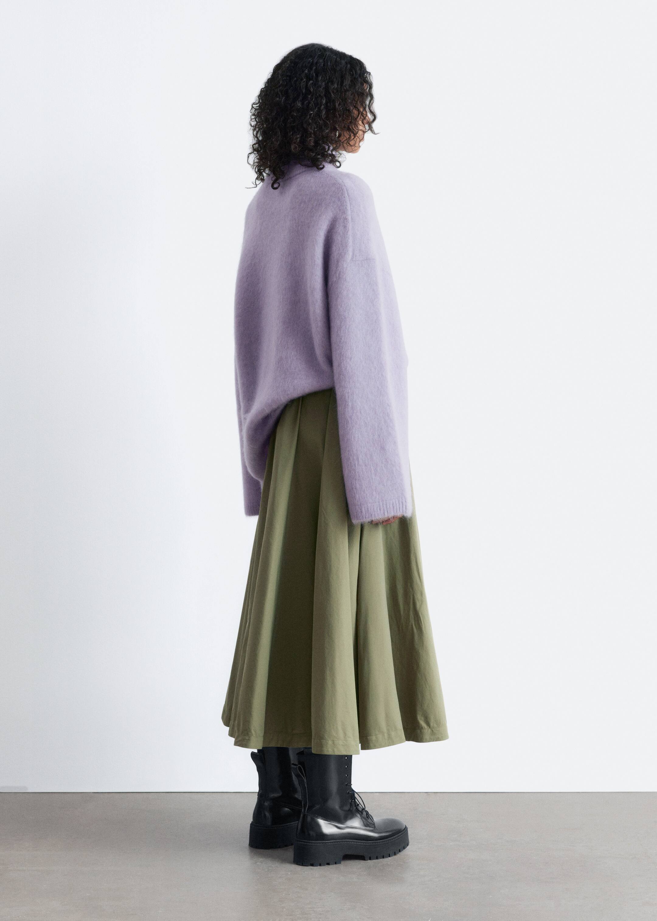 Größeres Bild ansehen: Ein olivgrüner Midirock, der durch eine weite A-Linien-Silhouette und sanfte Falten auffällt, wird von dem Model getragen. Es wird mit einem lavendelfarbenen Pullover und schwarzen Stiefeln kombiniert.