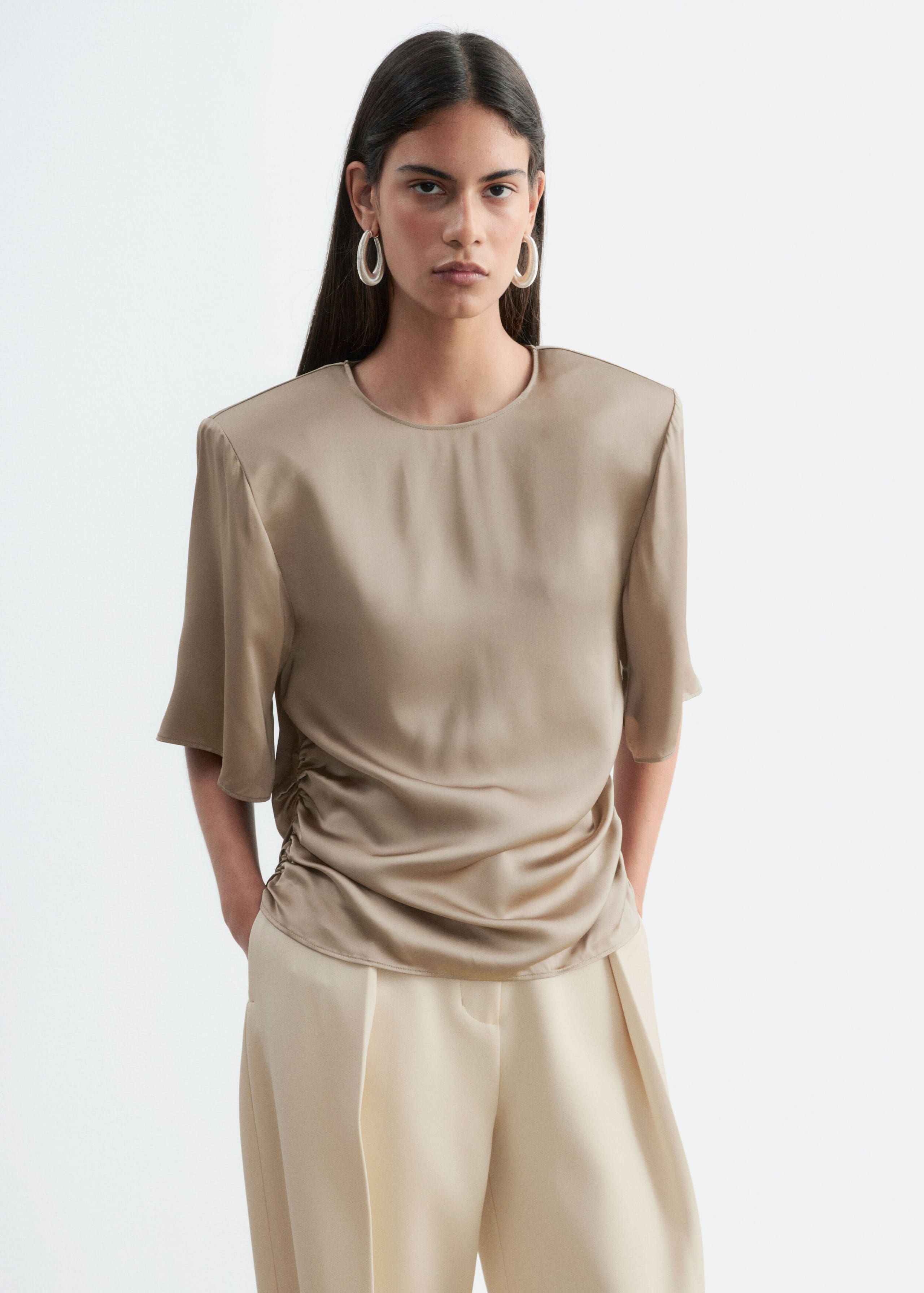 The model is wearing a taupe satin blouse with structured shoulder pads, short wide sleeves, and gathered side pleats, styled with cream-colored trousers.