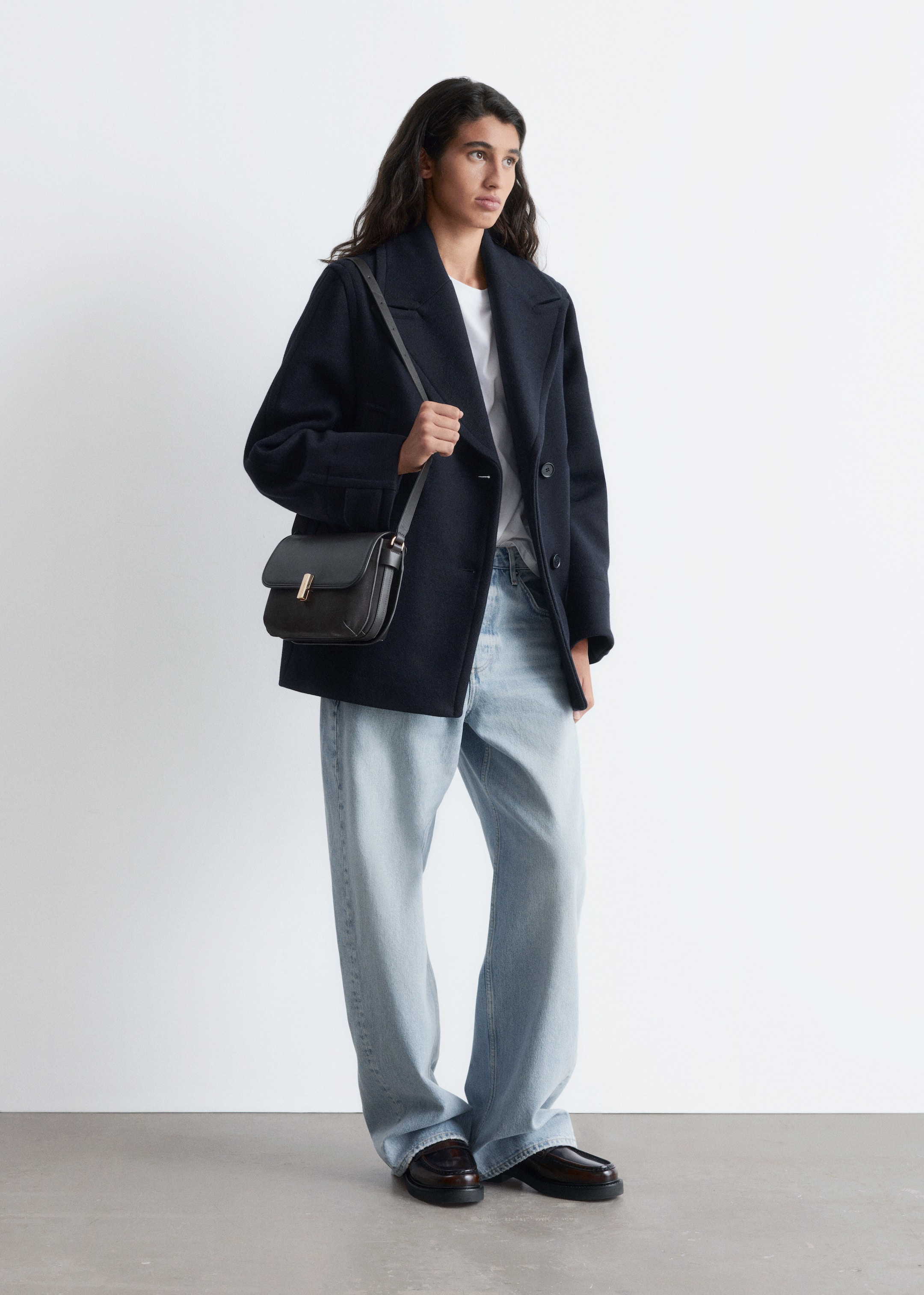 View larger image: A dark blue peacoat is being presented from a front-right angle, featuring wide notch lapels and a double-breasted front. It is showcasing a relaxed, boxy fit with two-button tabs visible on the cuffs.