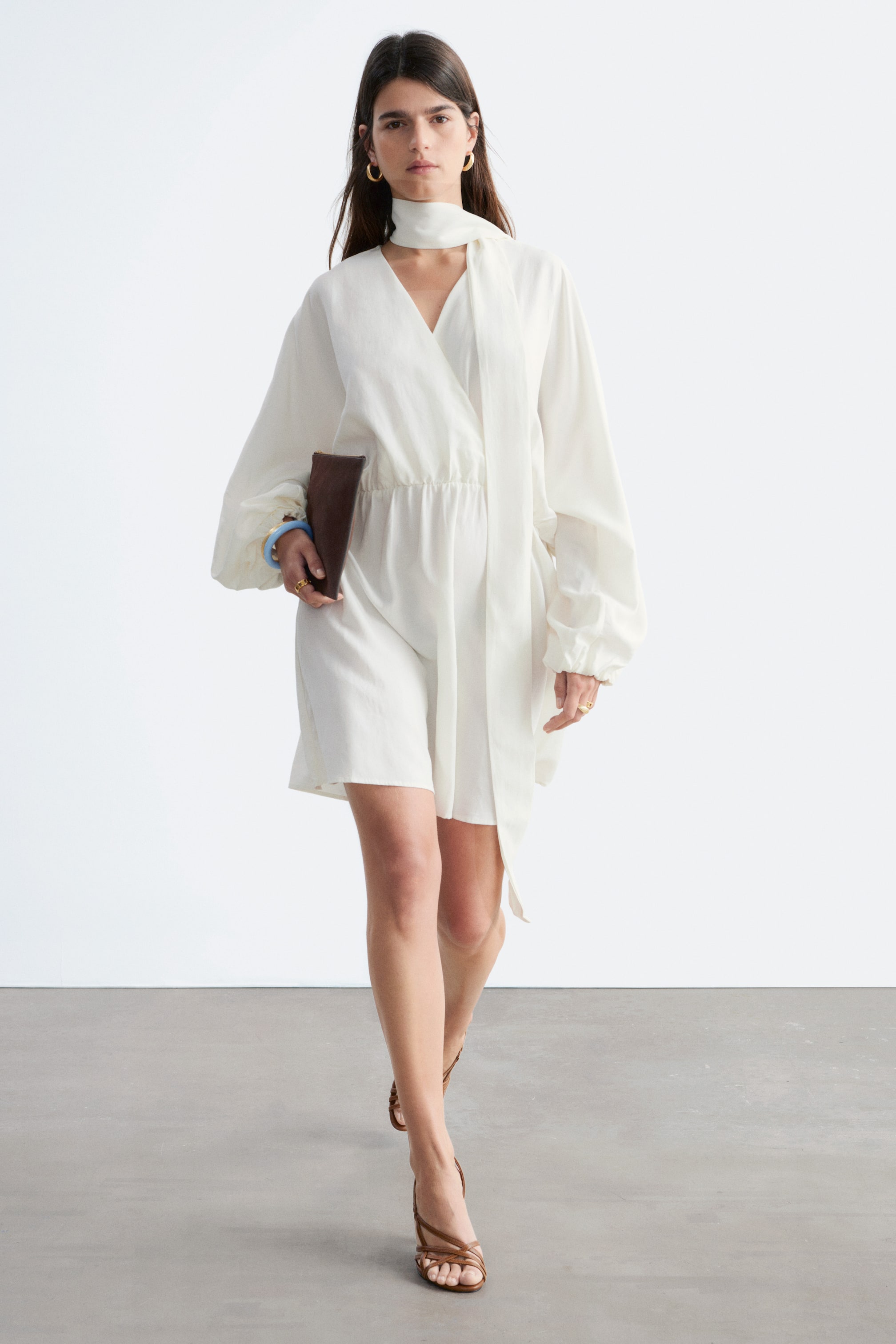 Grotere afbeelding bekijken: Een witte playsuit met een overslag V-hals en een geïntegreerde lange strikdetail rond de hals. Het ontwerp heeft volumineuze lange mouwen, elastische manchetten en een elastische tailleband die leidt naar loszittende shorts.