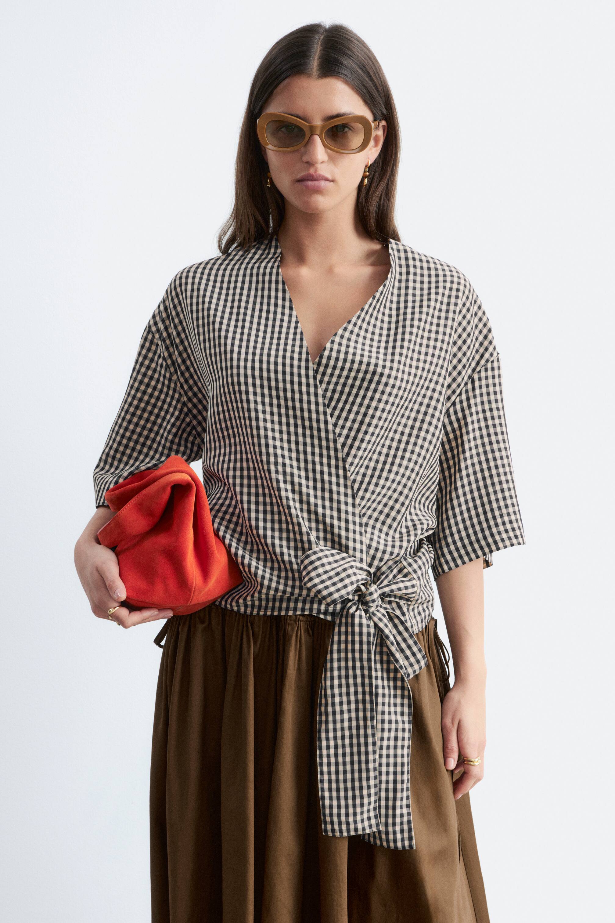 Ver imagem maior: A modelo está a usar uma blusa cruzada de padrão xadrez preto e branco, com decote em V e mangas curtas. A blusa está atada com um nó na cintura, e a modelo segura uma clutch vermelha enquanto usa também uma saia castanha.