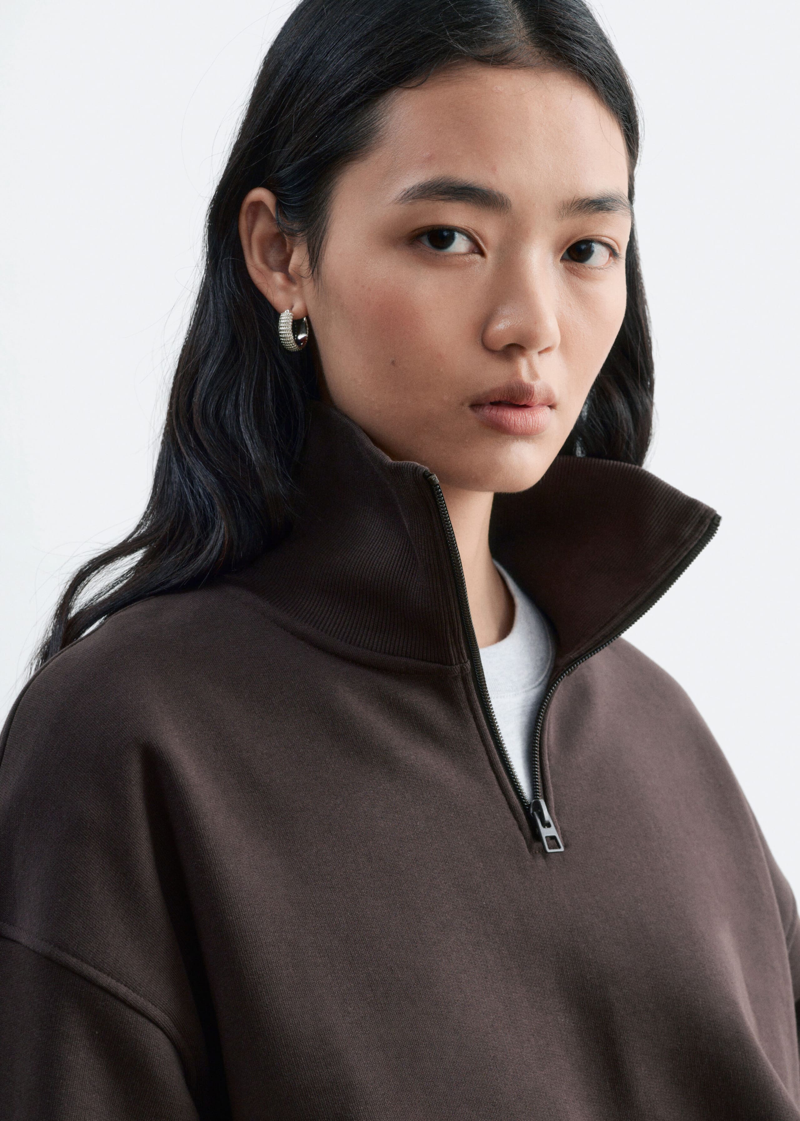 A dark brown cotton jersey sweatshirt featuring a half-zip neckline with a ribbed collar is being worn by the model. The zip is partially opened, revealing a light grey crew-neck T-shirt layered underneath.