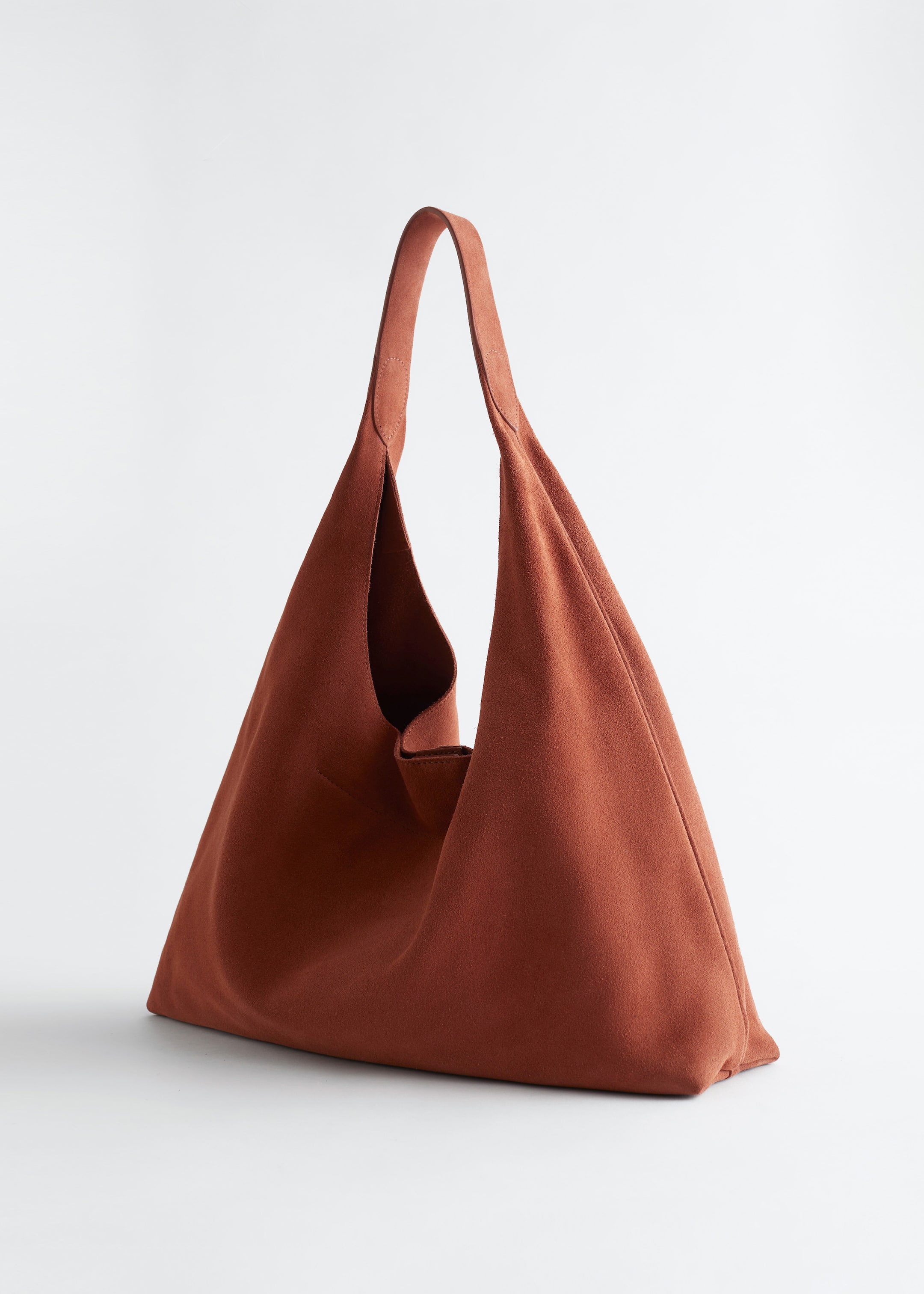 Ver imagem maior: Uma tote bag de cor ferrugem, confeccionada em camurça suave com acabamento mate, exibe a sua forma macia e descontraída. Apresenta uma alça larga única e uma costura decorativa vertical no centro da frente.