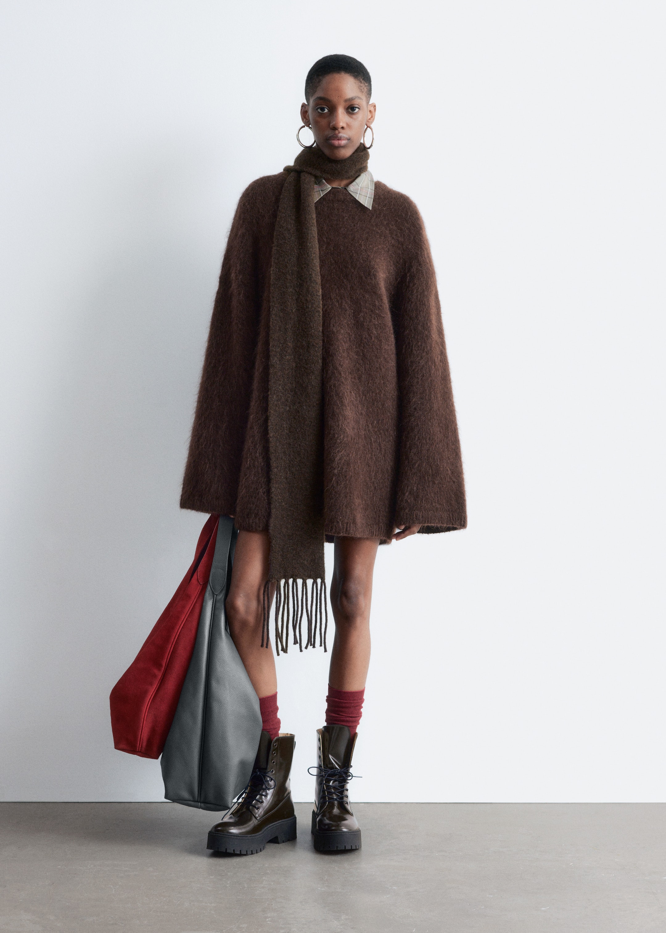 View larger image: A brown, shaggy oversized sweater is being presented from a full-front view, layered over a light plaid collar, with a dark brown fringed scarf draped around the neck. Dark, glossy combat boots with thick lug soles are being worn, and two large tote bags, one maroon and one grey, are being held.