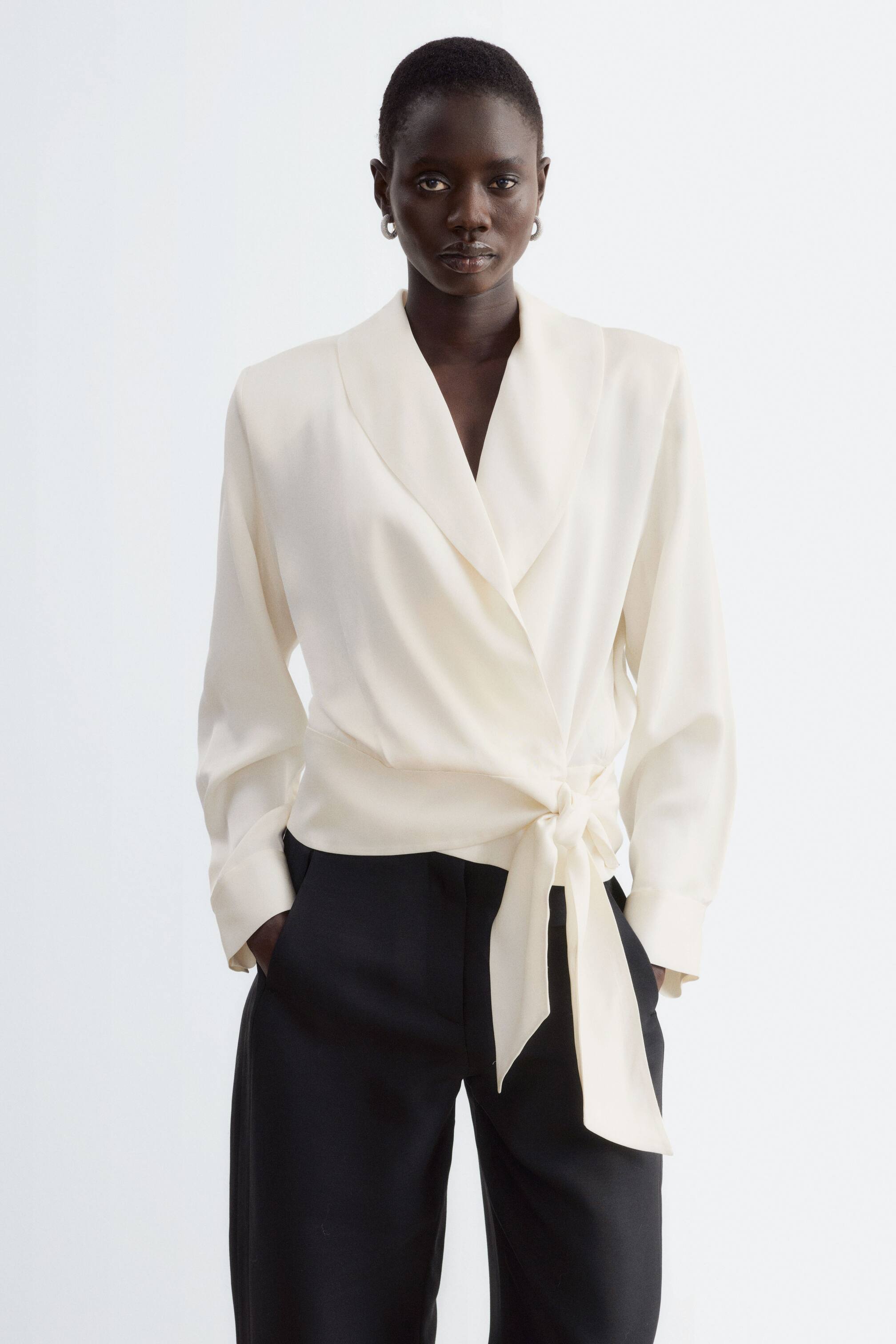 Visualizza l'immagine ingrandita: La modella indossa una blusa avvolgente color bianco sporco, con colletto a scialle, maniche lunghe e cintura laterale annodata sul davanti in vita. La blusa è abbinata a pantaloni neri.