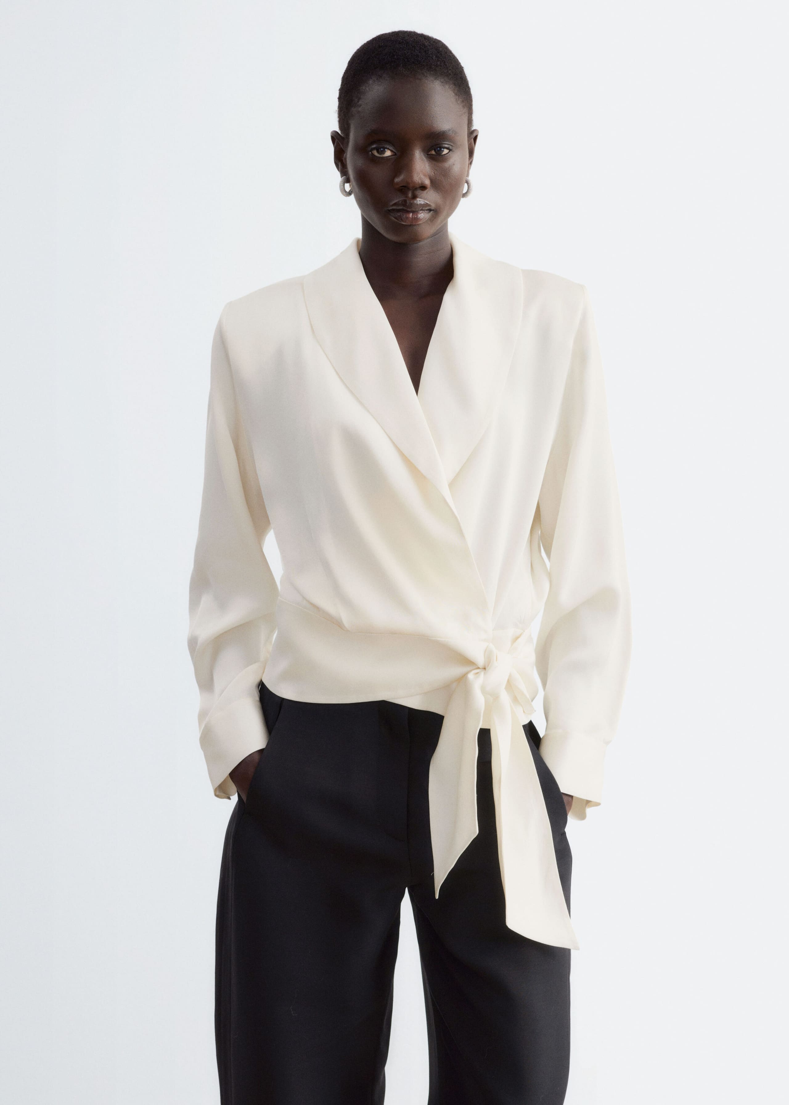 La modella indossa una blusa avvolgente color bianco sporco con colletto a scialle, maniche lunghe e un laccio laterale sul davanti in vita. La blusa è abbinata a pantaloni neri.