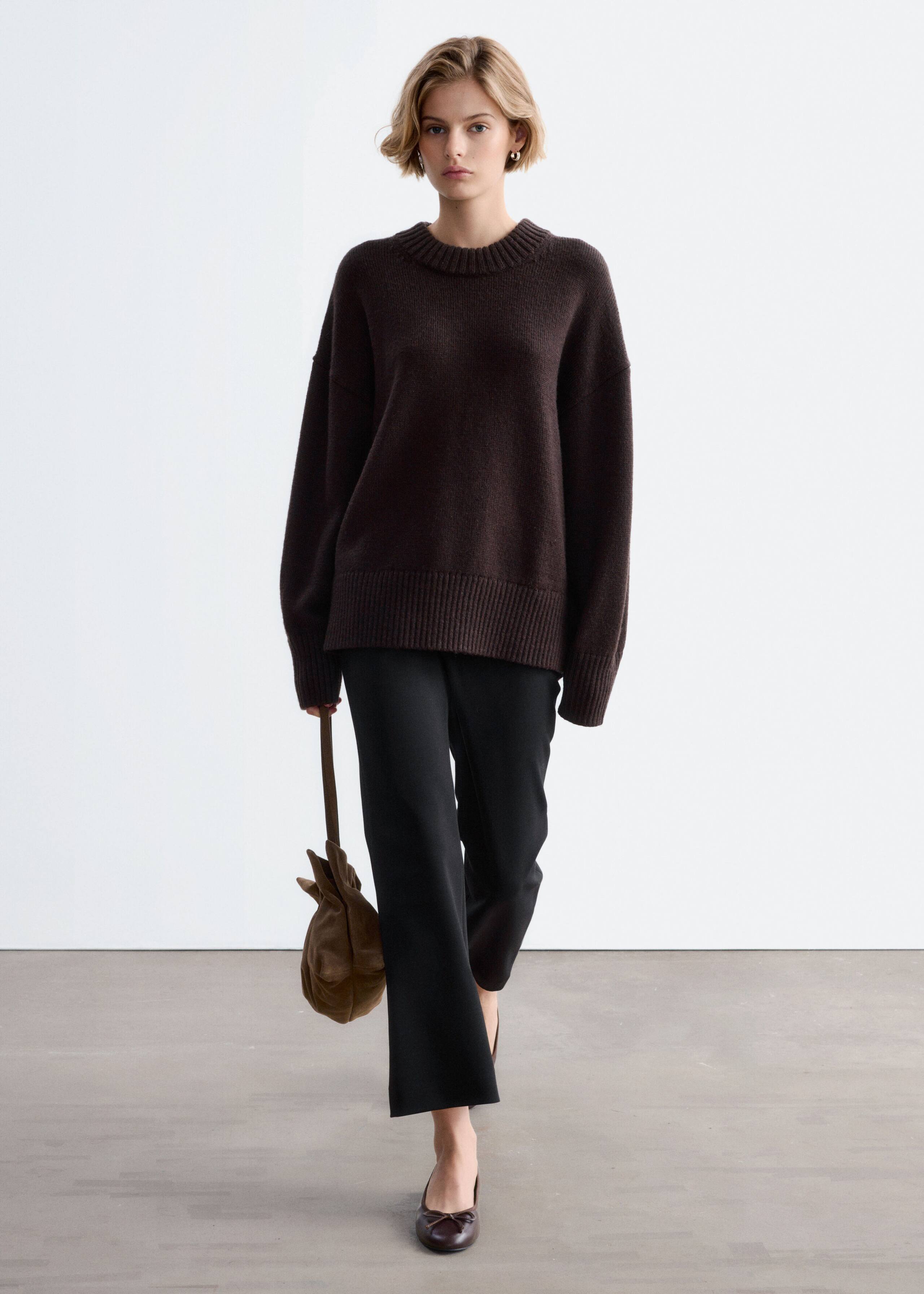 The model is wearing an oversized dark brown wool sweater featuring a crew neck, dropped shoulders, and ribbed detailing at the collar, cuffs, and hem. The sweater is styled with black straight-leg trousers and a brown suede handbag held in one hand.