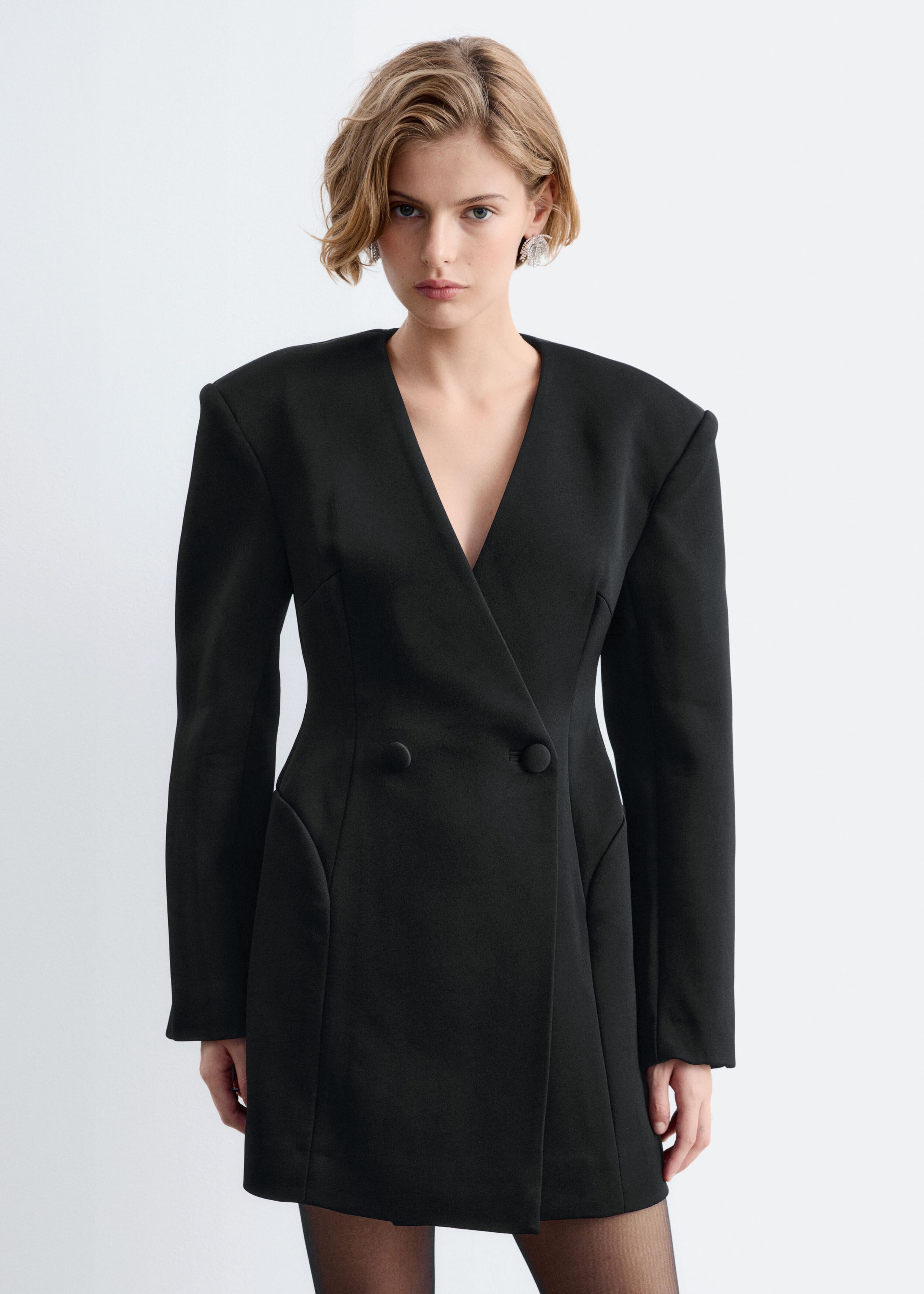 A black double-breasted blazer dress with structured shoulders and a cinched waist is worn by the model. The collarless dress features a deep V-neckline, vertical seam details, and two fabric-covered buttons at the front.
